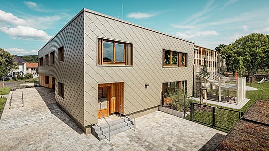 Das Bild zeigt die kommunale Kindertagesstätte in Alzenau aus einer schrägen Vogelperspektive und betont die moderne Architektur mit ihrer markanten PREFA Aluminiumfassade in Bronze. Die Fassade besteht aus Wandrauten 44 × 44, die ein harmonisches Schuppenmuster erzeugen und dem Baukörper eine elegante Dynamik verleihen. Die großen Holzfenster mit vertikalen Lamellen setzen warme Akzente und lassen viel Tageslicht ins Innere. Rechts im Bild öffnet sich das Gelände mit einem eingezäunten Spielbereich, Grünflächen und einer überdachten Terrasse. Die Kombination aus langlebigem, wartungsfreiem Aluminium und nachhaltigen Materialien macht das Gebäude zum Vorzeigeprojekt für funktionale und ökologische Architektur im Bildungsbau.
