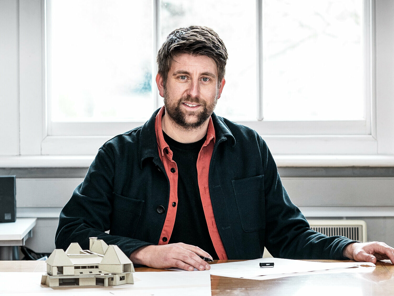 Portrait of the lead architect Tim Howell of the British office Mitchell Evans Architects sitting in front of a model at a table.