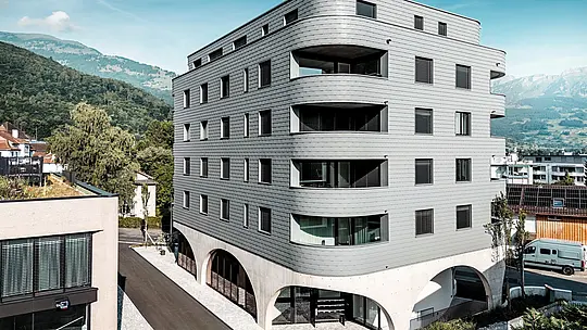 Außenansicht des Bernerhauses in Buchs mit markanter, dreieckiger Form und weich gerundeten Balkonbändern. Die Fassade aus PREFA Wandschindeln in P.10 Hellgrau umhüllt das Gebäude wie eine schuppenartige Aluminiumhaut und erzeugt ein präzises, lebendiges Fassadenbild. Über dem massiven Betonsockel mit eleganten Arkaden wirkt die leichte, witterungsbeständige PREFA Aluminiumfassade besonders ausdrucksstark. Langlebig, wartungsarm und formstabil setzt sie vor alpiner Bergkulisse ein modernes architektonisches Statement.
