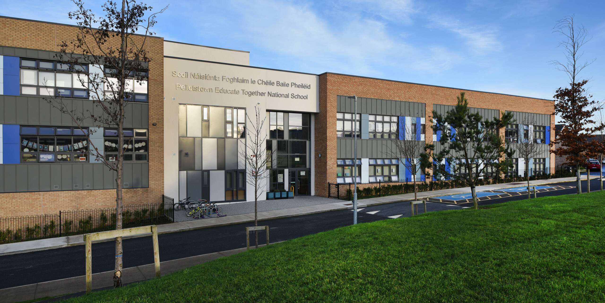 Front view of the Pelletstown Educate Together National School in Dublin with modern PREFALZ façade in P.10 light grey. The harmonious combination of aluminium, brick and plaster underlines the durable and elegant design of the school building. A well-tended green area with trees in the foreground emphasises the commitment to sustainability - as do the bicycles in front of the entrance.