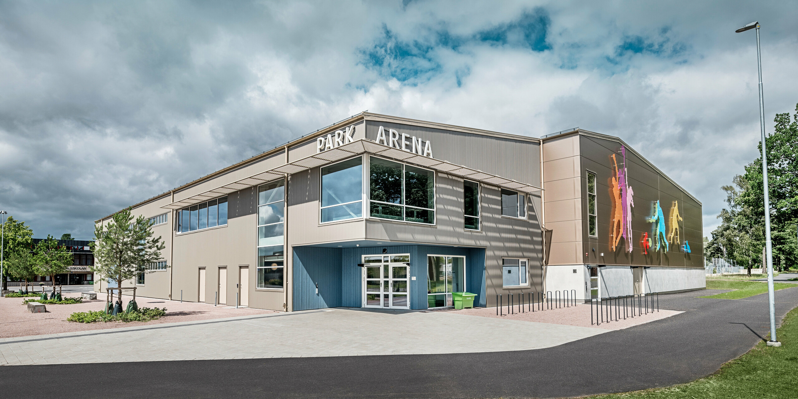 Frontansicht der Park Arena in Värnamo, Schweden, mit moderner PREFA Fassade aus Zackenprofilen in Bronze. Die Aluminiumfassade bietet eine langlebige und wetterbeständige Lösung, die durch markante Licht- und Schatteneffekte hervorsticht. Ein künstlerisch gestaltetes Wandbild auf der rechten Seite zeigt farbenfrohe sportliche Figuren, die den dynamischen Charakter der Sporthalle betonen. Große Fensterflächen lassen viel natürliches Licht in das Gebäude und verstärken das moderne Design der Arena.