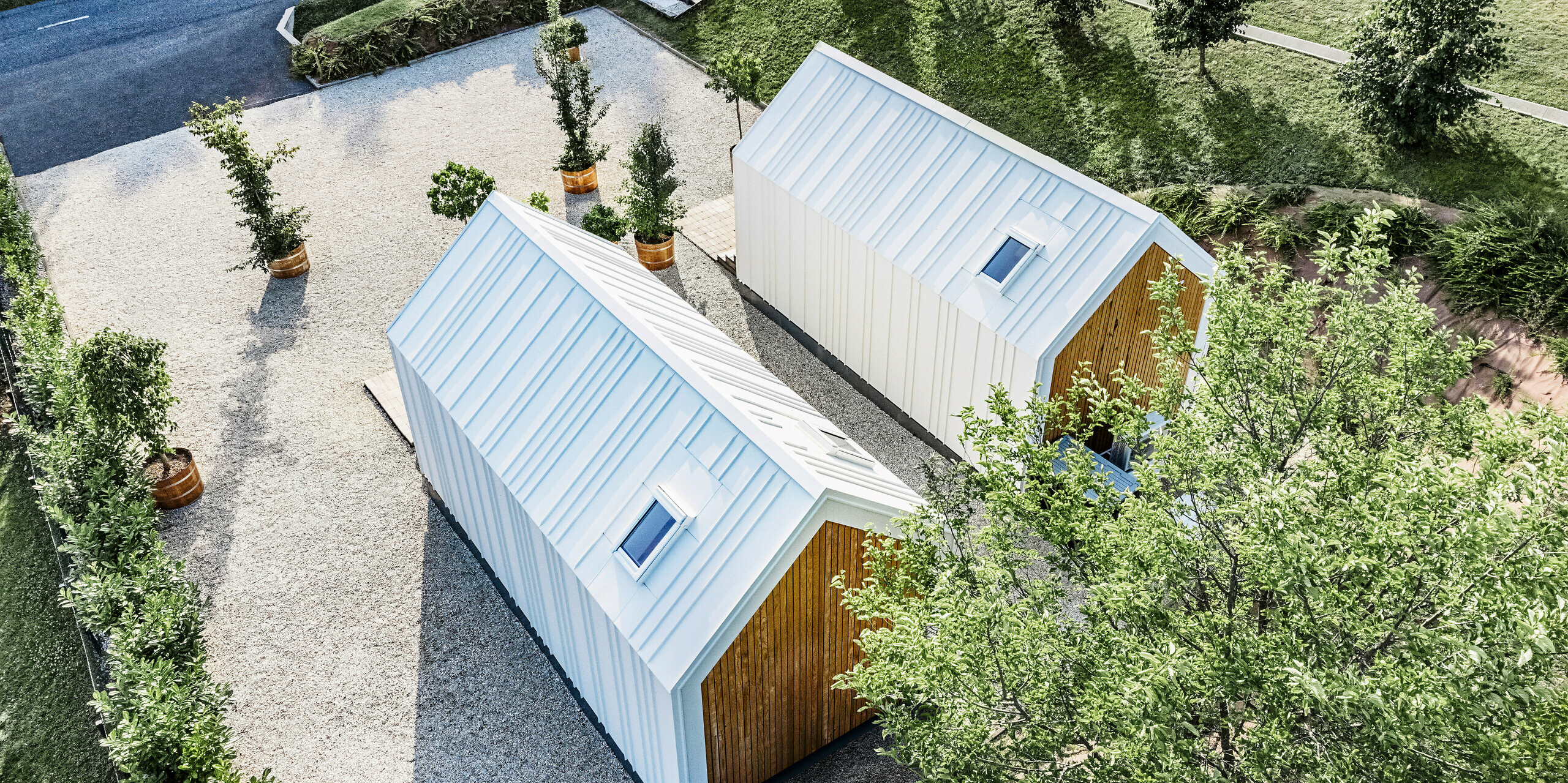 Bird's eye view of the rear of the two tiny houses 'Pri Momi' in Kančevci, Slovenia. The exclusive micro houses are equipped with robust PREFALZ roof and façade cladding in the colour P.10 pure white and natural wood. The white building envelope made of high-quality and durable sheet metal was designed with standing seam cladding. The minimalist architecture blends harmoniously into the surrounding green area and paved paths. The picture also shows the slightly staggered arrangement of the buildings, which were erected using timber frame construction.