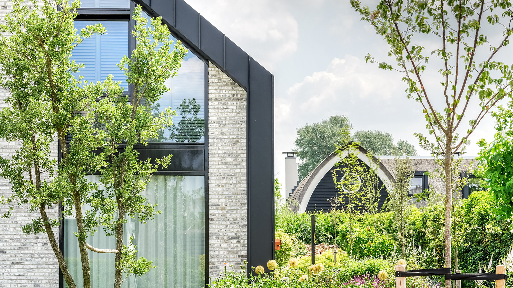 A modern detached house with a black Prefalz roof, nestled in a green garden. The façade combines light-coloured bricks with large windows and dark frames. Another house with a similar roof style is visible in the background. The garden in the foreground is rich in a wide variety of plants and trees, which lend the picture a lively and natural atmosphere.