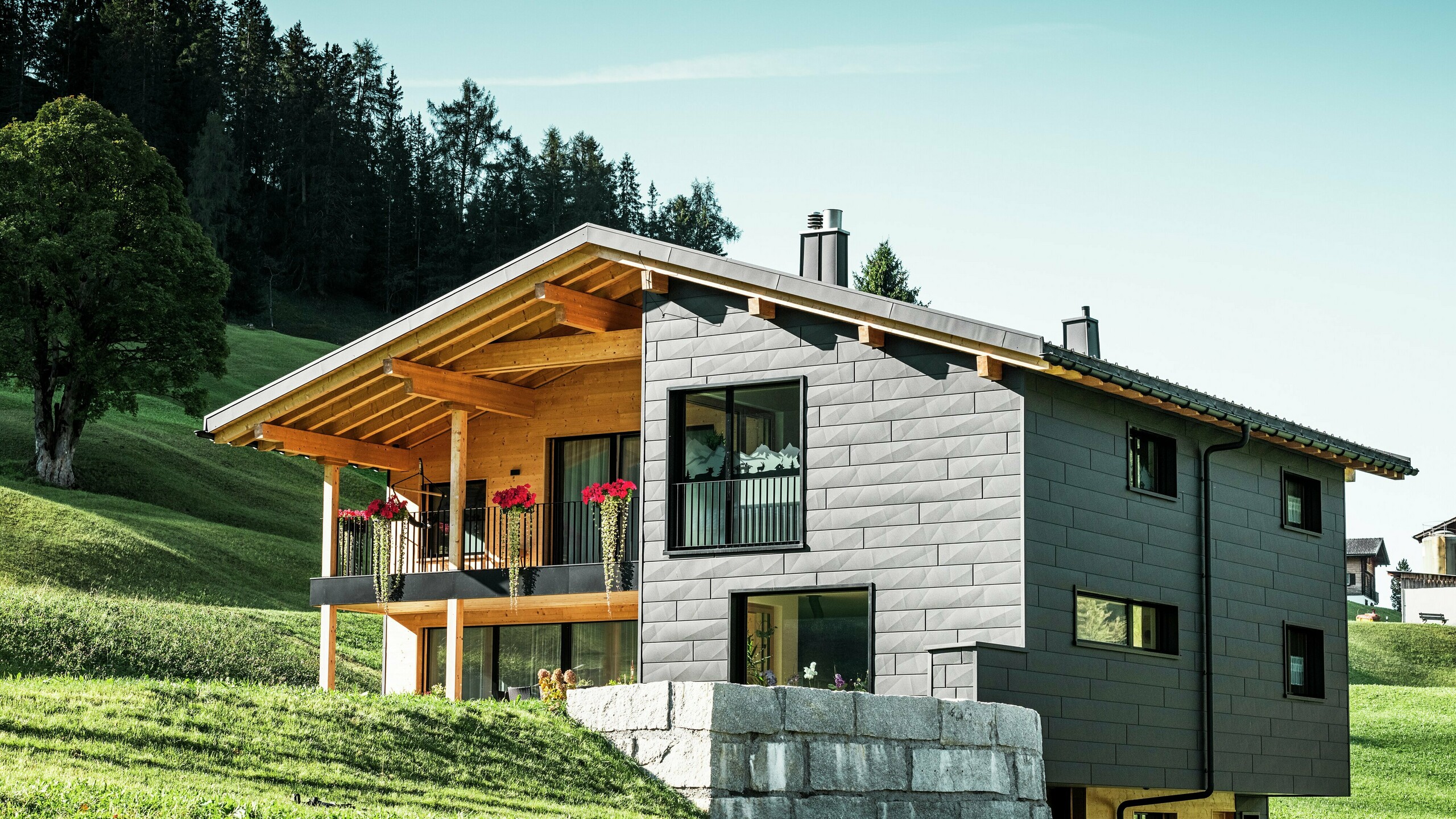 Dieses moderne Einfamilienhaus in Ascharina, Schweiz, fügt sich harmonisch in die grüne Alpenlandschaft ein. Das Gebäude ist komplett mit den langlebigen und witterungsbeständigen PREFA Siding.X Fassadenpaneelen in P.10 Schwarz verkleidet, die dem Bauwerk ein einzigartiges Erscheinungsbild verleihen. Der Holzbalkon ergänzt die dunkle Aluminiumhülle perfekt und setzt einen natürlichen Akzent.