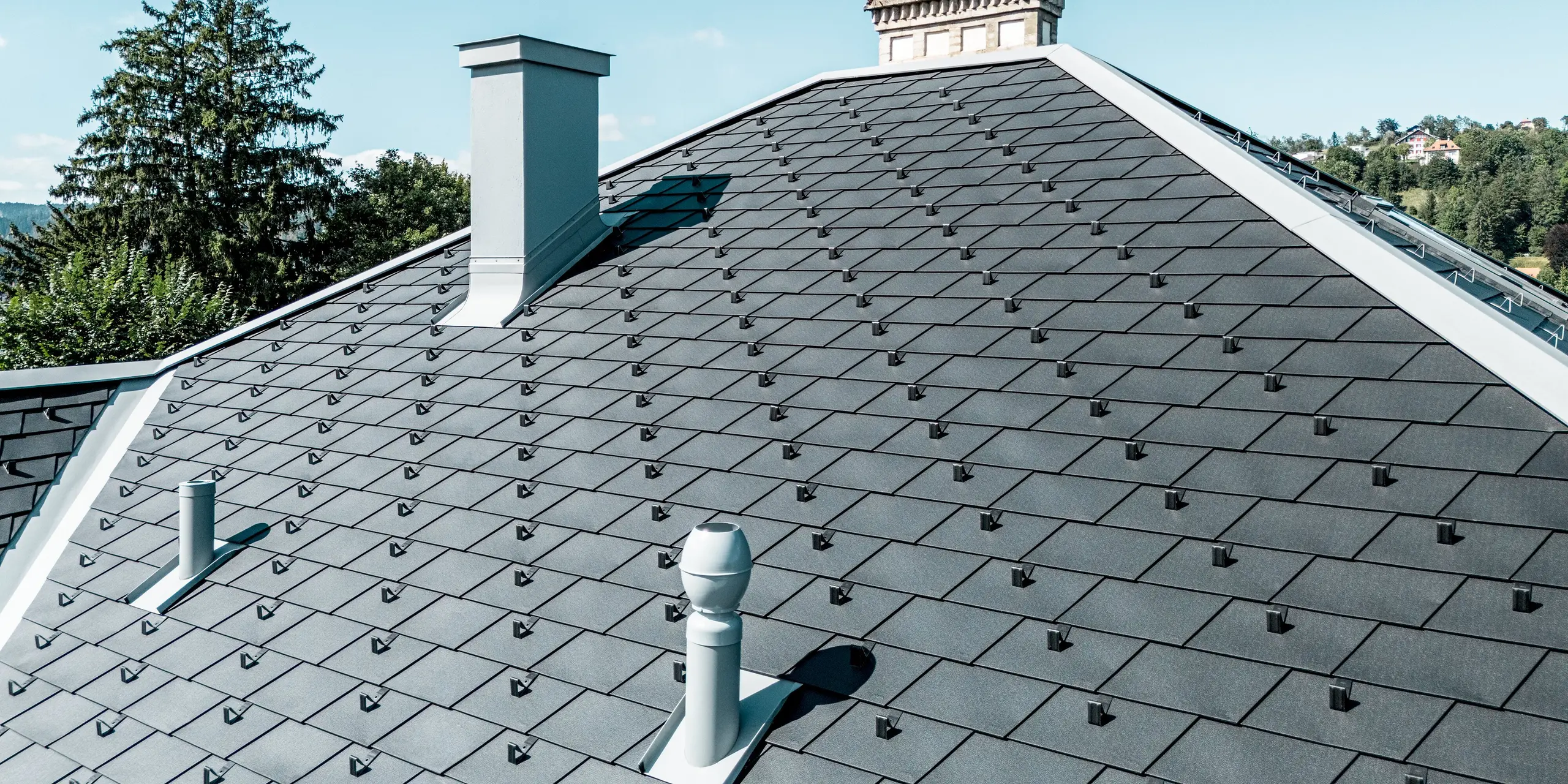 Detailaufnahme der Dachlandschaft des Manufacture Horlogère du Locle in der Schweiz: Die elegant geneigten Flächen sind mit PREFA Dachschindeln in P.10 Schwarz gedeckt und ziehen mit ihren präzisen Linien sofort den Blick an. PREFALZ in Patinagrau setzt moderne Akzente an Übergängen, Gauben und Kanten. Das leichte, langlebige Aluminiumdach überzeugt durch hohe Widerstandsfähigkeit, flexible Verarbeitung bei komplexen Formen und sorgt für ein harmonisches Zusammenspiel aus architektonischer Raffinesse und dauerhafter Qualität.