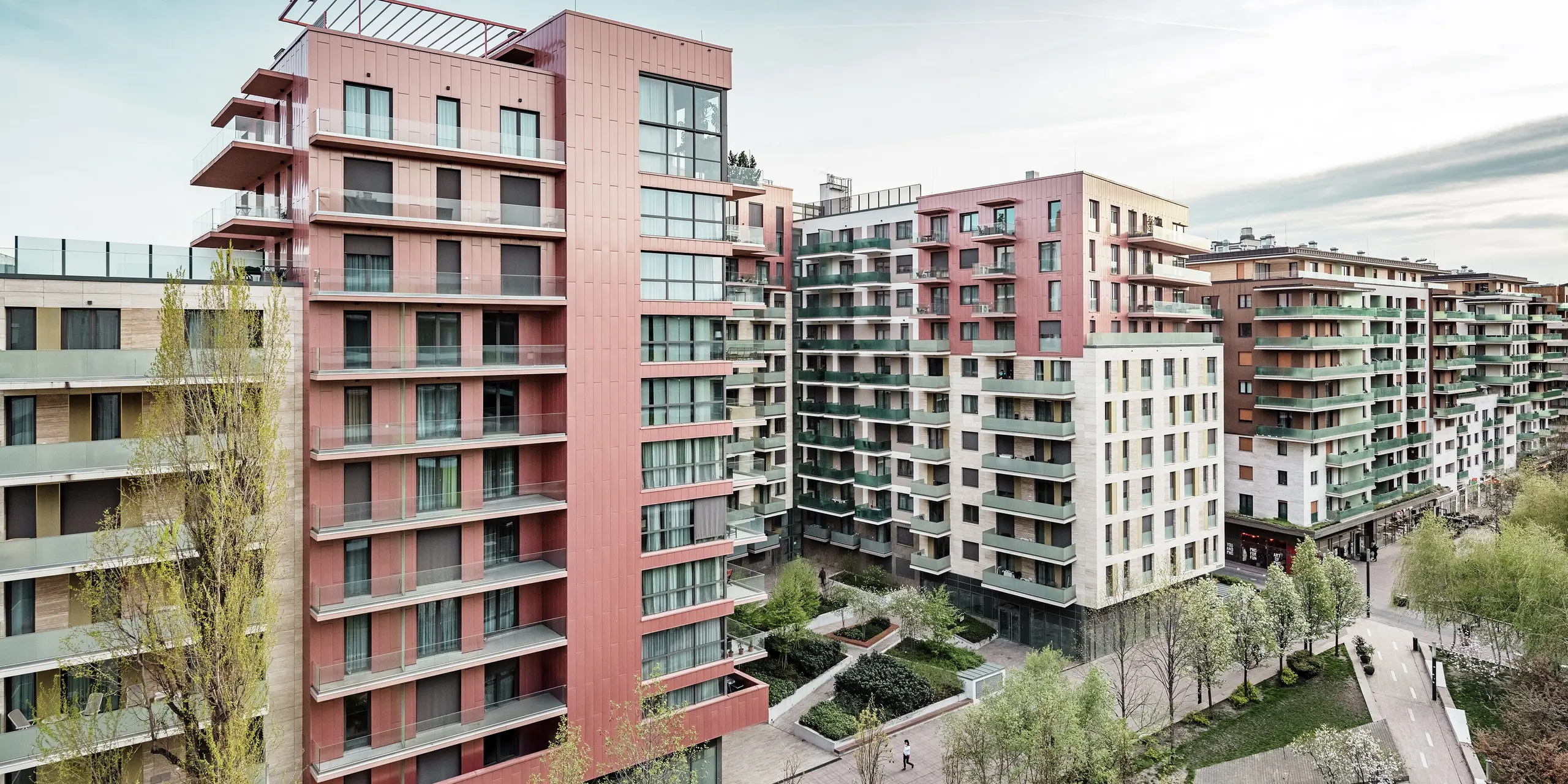 Panoramaaufnahme des Grand Corvin Wohnkomplexes im neugestalteten Corvin-Viertels in Budapest. Die markanten Gebäudeteile sind mit über 800 m² PREFA Siding Fassadenpaneelen in vertikaler Verlegung und Sonderfarbe Lachsmetallic versehen. Die langlebige Aluminiumverkleidung setzt architektonische Highlights, fügt sich harmonisch in die urbane Struktur ein und unterstreicht den hochwertigen, zeitgemäßen Charakter der neuen Wohnanlage entlang der Corvin-Promenade.