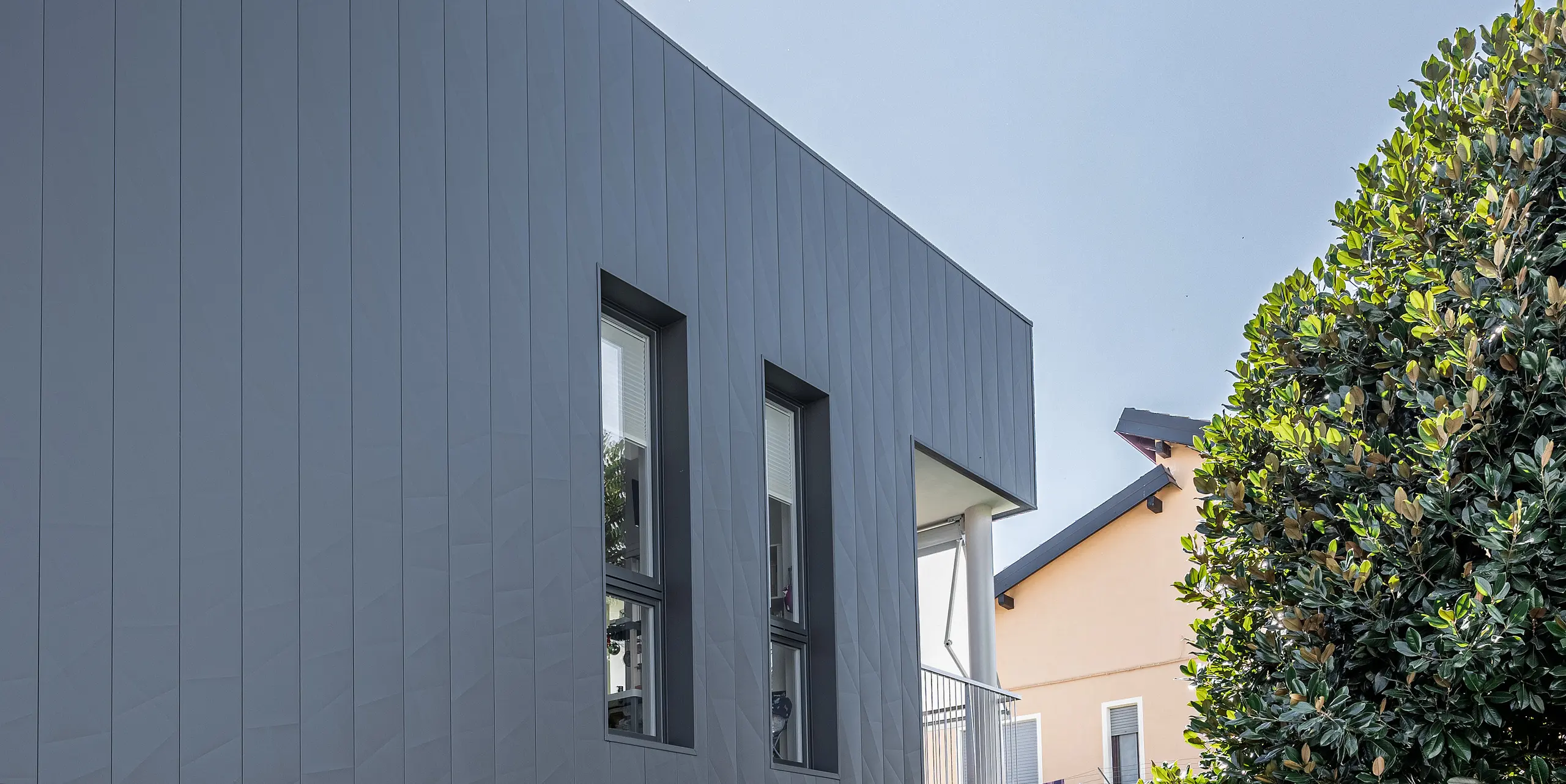 ertikale Ansicht eines zeitgenössischen Einfamilienhauses in Savigliano, Italien, ausgestattet mit einer dunkelgrauen PREFA Siding.X Aluminiumfassade. Die Fassade aus Blech zeichnet sich durch ihre einzigartige matte Oberfläche und geometrische Struktur aus, die das moderne Design des Hauses betont. Großflächige Glasfronten bieten einen transparenten Übergang zwischen dem Innenraum und dem sorgfältig angelegten Garten, wodurch eine offene und einladende Wohnatmosphäre entsteht.