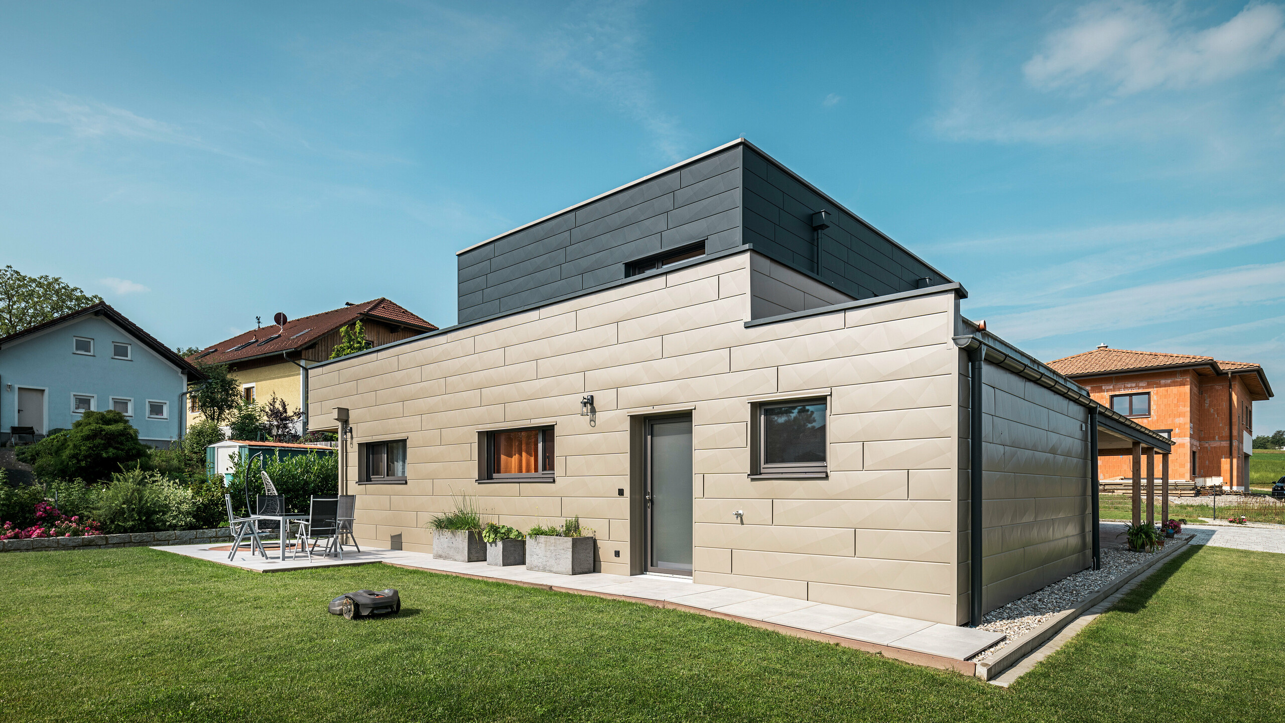 Modernes Einfamilienhaus in Wippenham, Österreich, mit zweifarbiger PREFA Aluminiumfassade in Bronze und Anthrazit. Die Fassade ist mit Siding.X Fassadenpaneelen gestaltet, die für eine elegante und zeitgemäße Optik sorgen. Die integrierte PREFA Dachentwässerung mit Wasserfangkasten und Ablaufrohr sowie eine gepflegte Gartenanlage mit Terrasse betonen das moderne Design des Hauses.