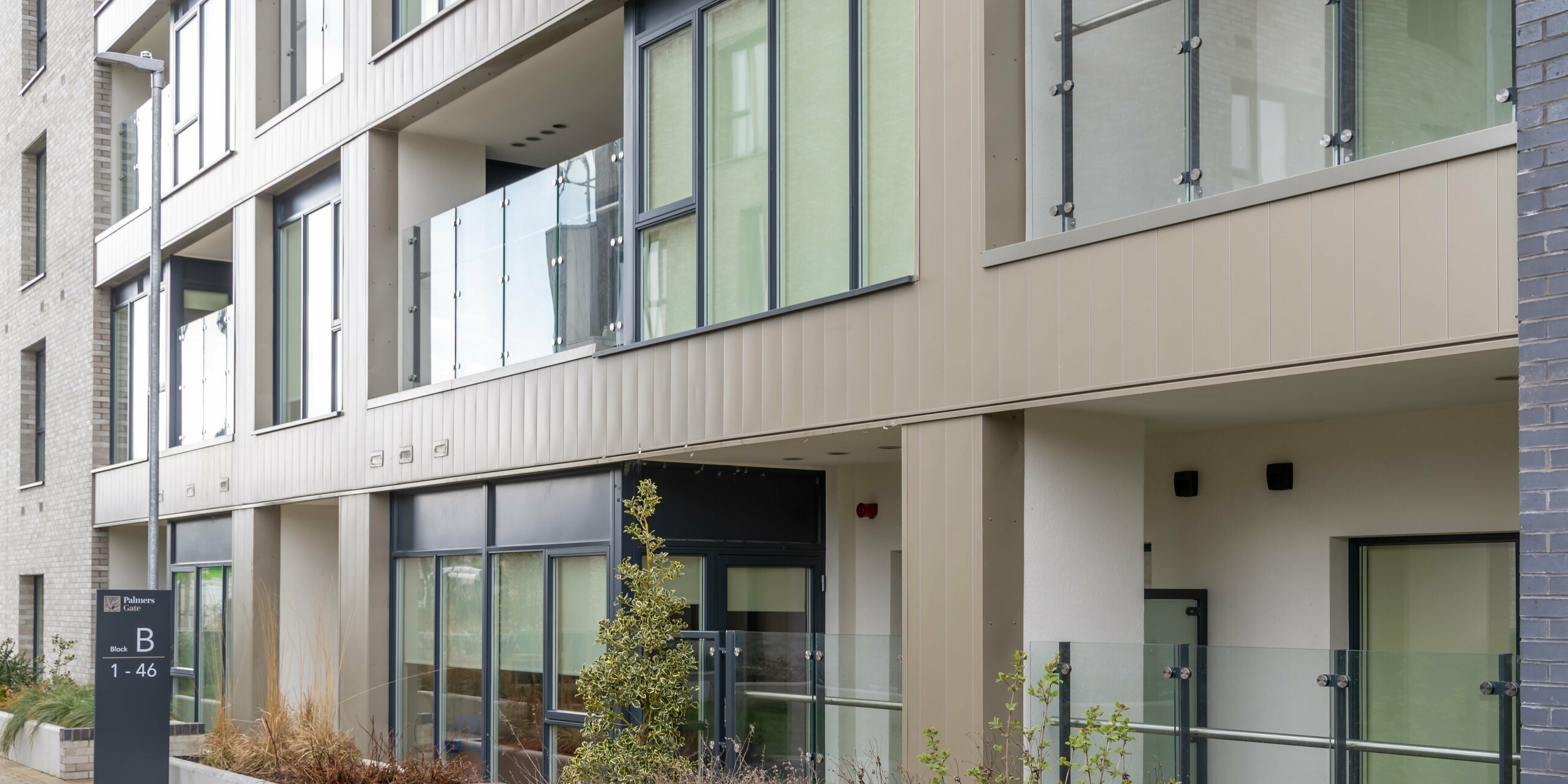 Entrance area of apartment block B in the Palmers Gate residential complex in Palmerstown, Dublin, Ireland. The façade is clad with bronze-coloured PREFA sidings and the PREFALZ façade system, giving it a modern and elegant appearance. Glass balustrades on the balconies and planters in front of the windows give the building additional charm. The path to the entrance is paved with cobblestones and lined with green plants. The architectural details and the choice of high quality materials emphasise the functionality and aesthetics of the complex.