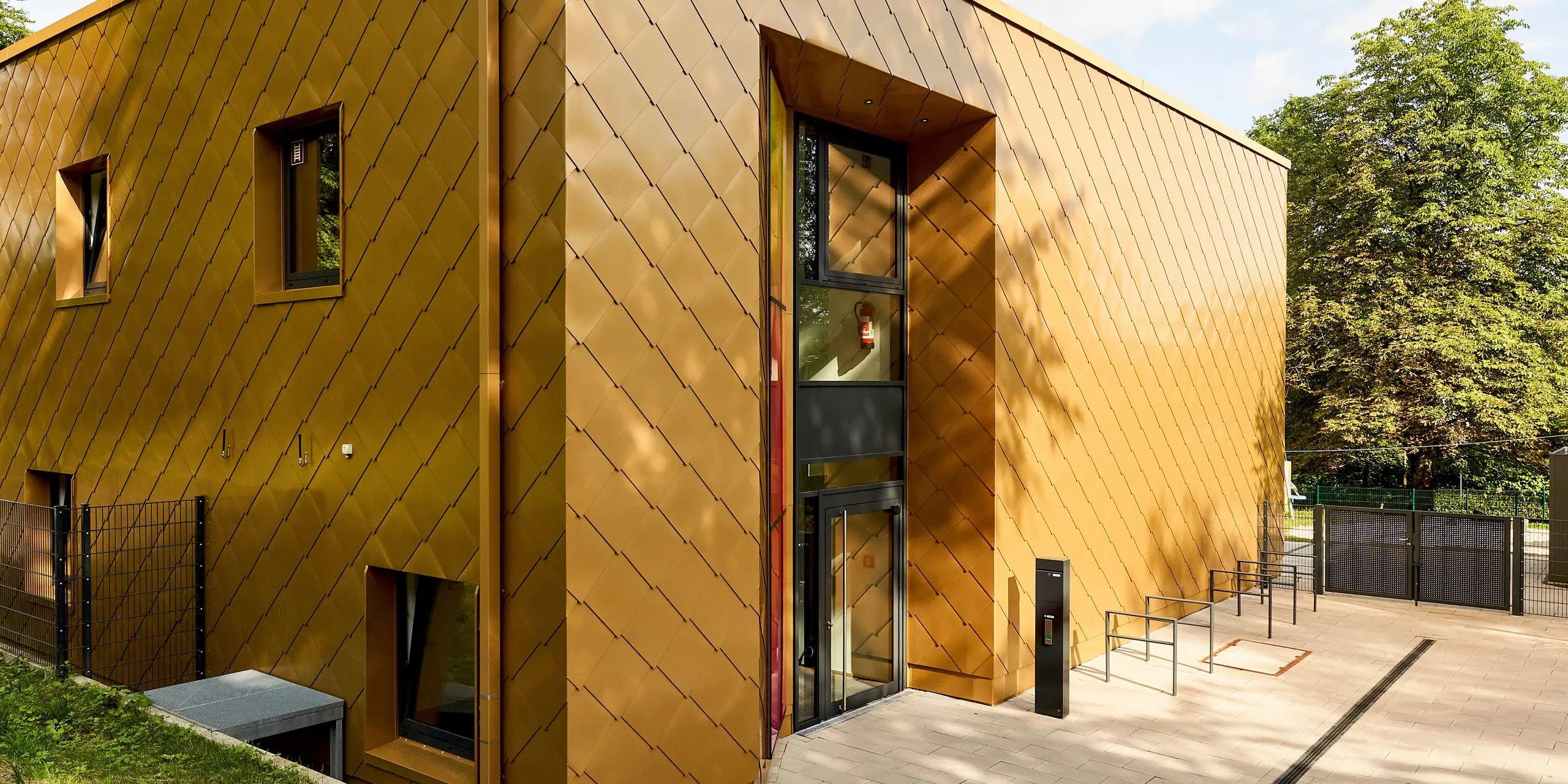 Moderne Kubus-Architektur mit markanter PREFA Aluminiumfassade in goldener Schuppenoptik zieht sofort alle Blicke auf sich. Die präzise verlegten Rauten erzeugen ein lebendiges Spiel aus Licht und Schatten. Großzügige Fensterflächen und der klare Eingangsbereich unterstreichen die hochwertige Gestaltung. Die langlebigen, korrosionsbeständigen PREFA Aluminiumprodukte überzeugen durch extreme Widerstandsfähigkeit, Farbbrillanz und Wartungsfreiheit – perfekt für nachhaltige, stilprägende Fassadenlösungen in naturnaher Umgebung.