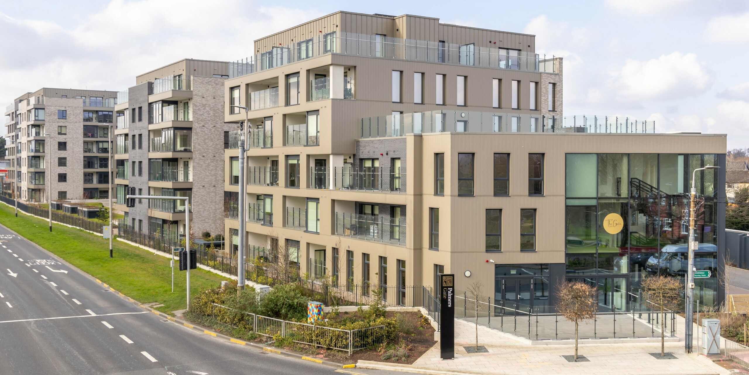 Exterior view of the Palmers Gate residential complex in Palmerstown, Dublin, Ireland. The complex comprises several residential buildings clad with bronze-coloured PREFA cladding and the PREFALZ façade system. The facades combine aluminium cladding with brick elements and large glass windows, emphasising the modern and elegant style. The buildings have balconies with glass balustrades and spacious communal areas. The entrance to the complex features large glass frontages and a 'Palmers Gate' sign. In total, approx. 1,700 m² of PREFA siding and approx. 1,200 m² of PREFALZ façade system were used.