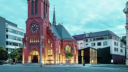 Das Bild zeigt die harmonische Fusion von historischer und moderner Architektur in Olmütz. Die beeindruckende Rote Kirche, mit ihrem markanten spitzen Turm und den gotischen Fenstern, bildet den historischen Mittelpunkt des Ensembles. Direkt daneben erhebt sich der moderne Neubau, dessen Fassade aus matten PREFABOND Aluminium-Verbundplatten und Glas einen klaren, eleganten Kontrast zur traditionellen Architektur bildet. Die großflächigen Glasfronten gewähren Einblicke in den Neubau, der auf innovative Weise mit der Kirche verbunden ist. Der moderne Bau trägt mit seiner präzisen Form und den klaren Linien zur zeitgenössischen Atmosphäre bei, ohne den historischen Charakter der Kirche zu beeinträchtigen. Diese gelungene Symbiose zeigt, wie hochwertig verarbeitete Materialien wie PREFABOND die perfekte Grundlage für die Verschmelzung von Tradition und Moderne bieten.