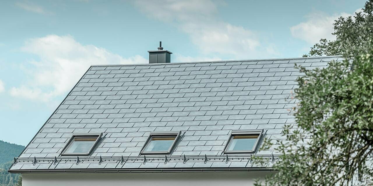 Classic gabled roof, clad in the PREFA R.16 roof tile in stone grey with three roof windows and snow guard system; the façade is rendered in white.