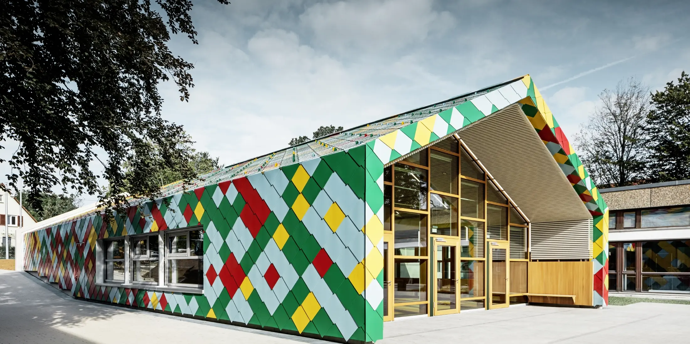 Modernes Gebäude mit auffälliger Fassade aus bunten Aluminiumrauten von PREFA in Grün, Rot, Gelb und Hellblau. Die Dach- und Wandflächen gehen fließend ineinander über und bilden ein geometrisches Muster. Der Eingangsbereich besteht aus großen Glasfronten mit Holzrahmen. Das Gebäude steht in einer grünen Umgebung mit Bäumen und weiteren Schul- oder Verwaltungsgebäuden im Hintergrund.