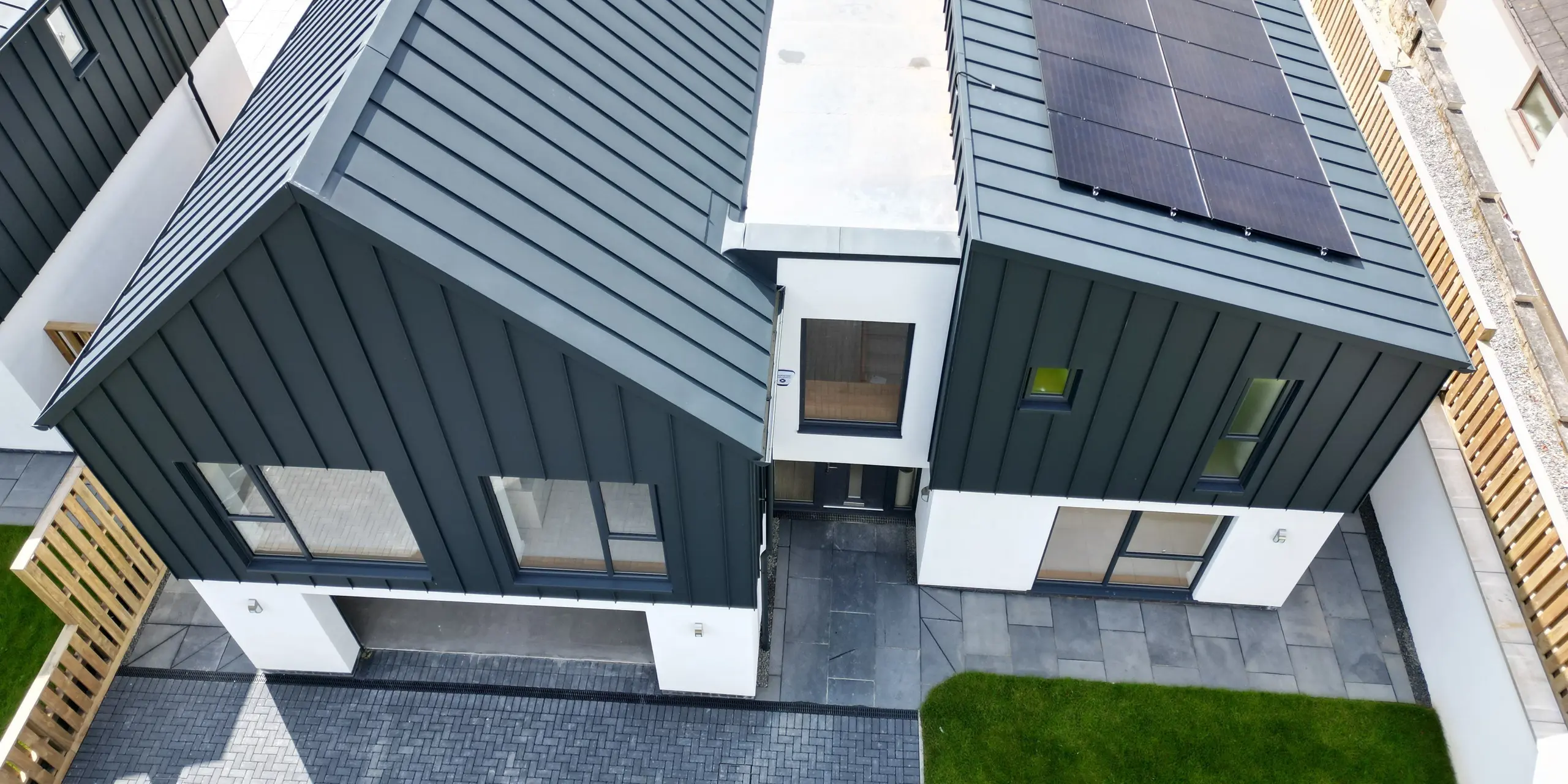 A bird's eye view of a detached house in Bridgerule, Devon, with PREFALZ P.10 anthracite as a standing seam covering on the roof and façade. The modern architecture combines durable aluminium with aesthetic longevity, complemented by a solar energy system. A gable roof towers over the garage entrance, while the right-hand part of the building has a pent roof. Between the roofs, a building bridge connects the two structures.
