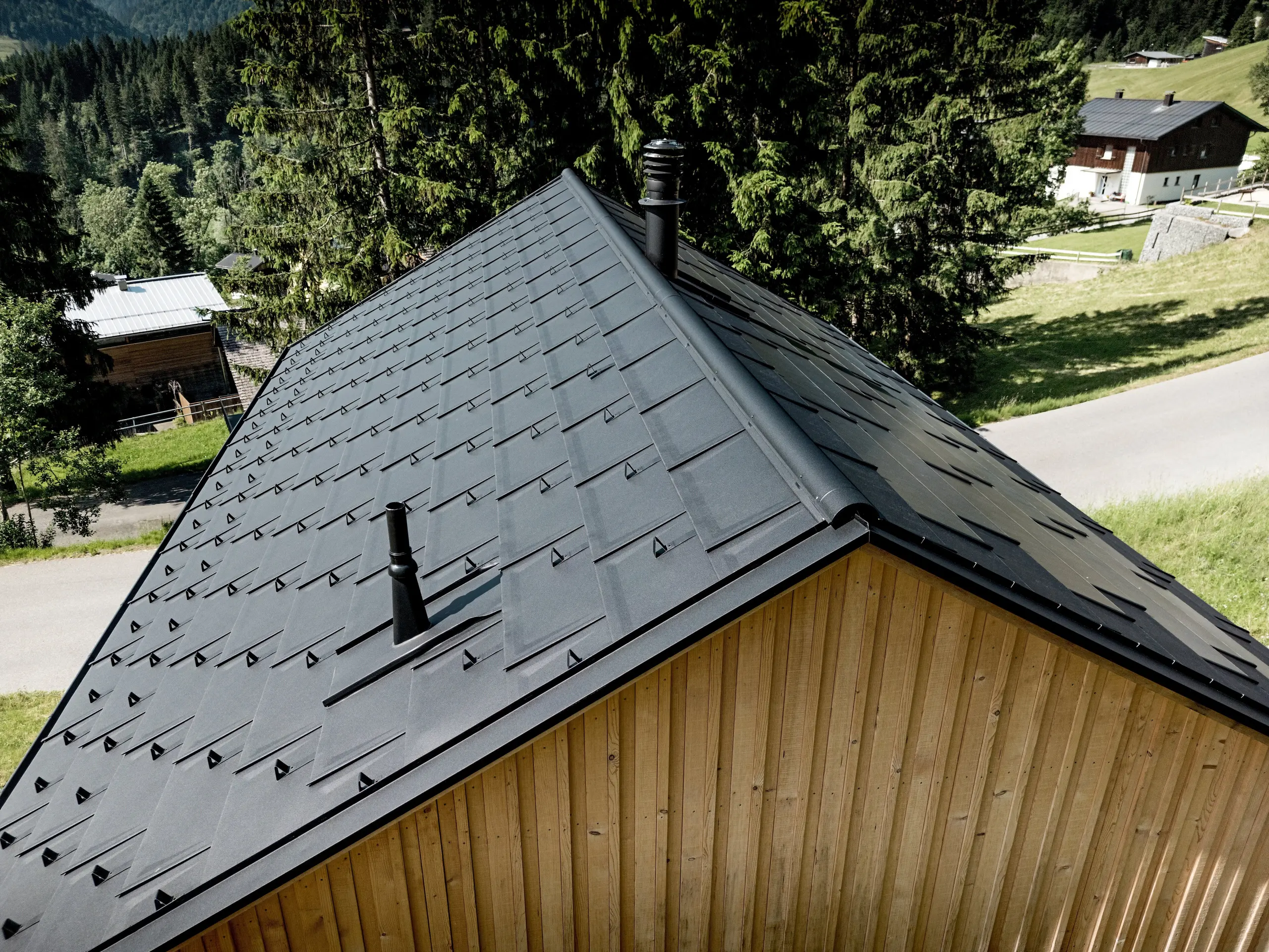 Die dynamische Nahaufnahme zeigt die markante Dachfläche von Haus Immi vor grünen Bergwiesen und dunklen Tannen. PREFA Dachplatten R.16 in P.10 Schwarz, präzise Detaillösungen und Schneefänger formen ein kraftvolles, einheitliches Dachbild. Die leichte, robuste Aluminiumdeckung widersteht alpinem Wetter sowie hohen Schneelasten und schützt das Holzhaus in Ebnit dauerhaft und wartungsarm.
