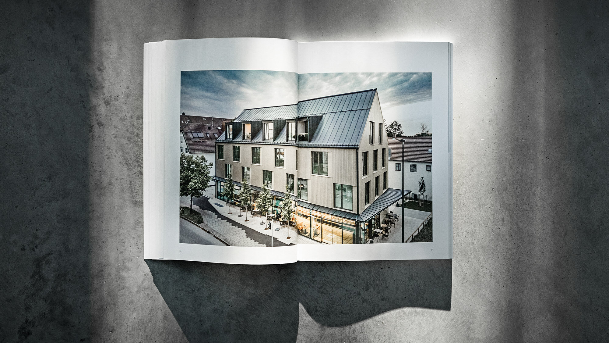 The open PREFARENZEN book 2024 with a double page showing the residential and commercial building Feldkirchen by Architekturbüro Heigl before a grey background.