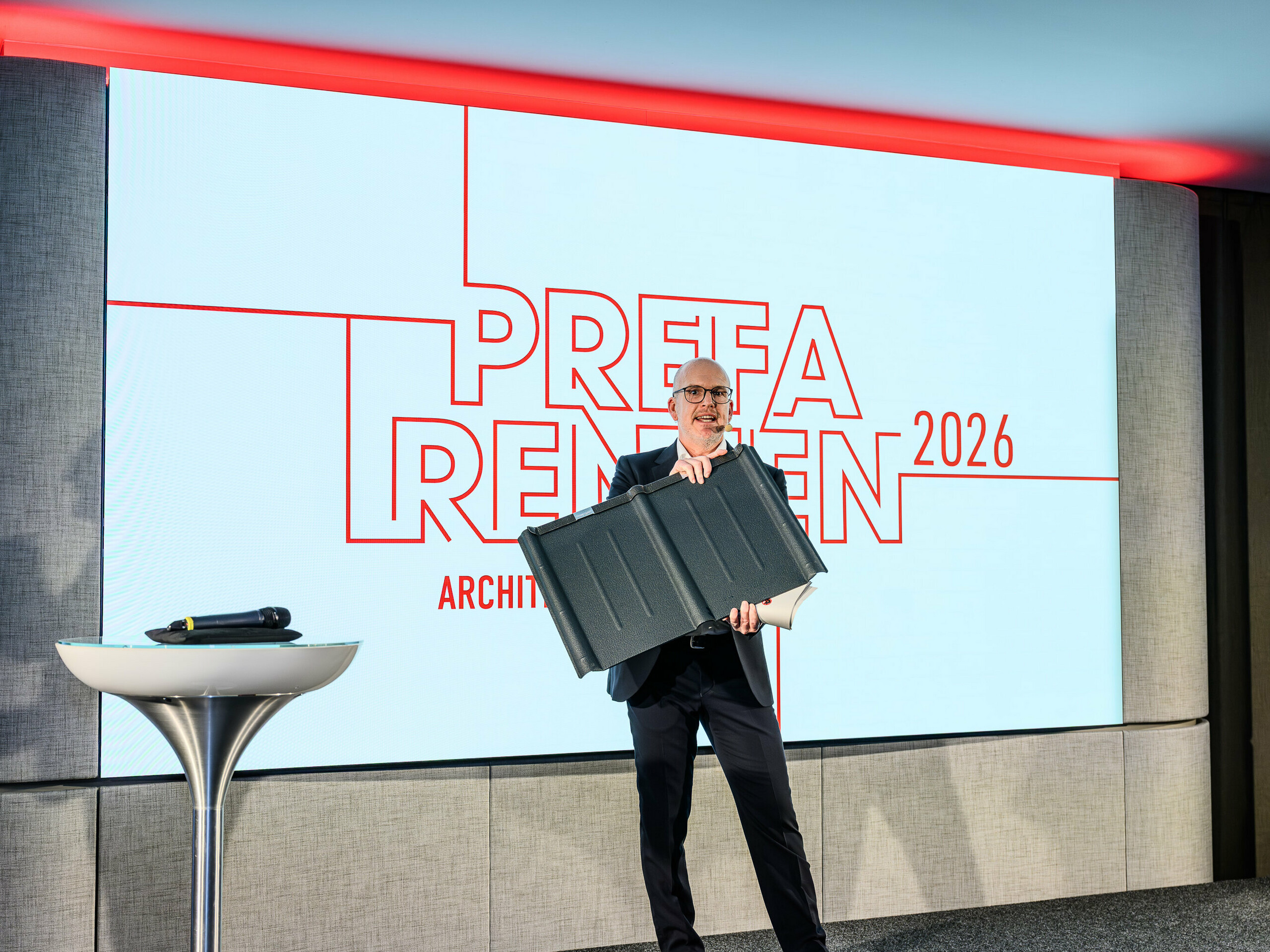 Highlight-Moment der PREFARENZEN Präsentation 2026: Jürgen Jungmair, PREFA Marketingleiter International, präsentiert stolz die legendäre PREFA Dachplatte. Im Fokus steht das langlebige Aluminiumprodukt, das seit rund 80 Jahren erfolgreich produziert wird. Die moderne Bühne mit dem markanten PREFARENZEN-Logo unterstreicht die Bedeutung des Produkts für zukunftsorientierte Architektur. Leicht, sturmsicher, rostfrei – die PREFA Dachplatte vereint Technik und Ästhetik auf höchstem Niveau.