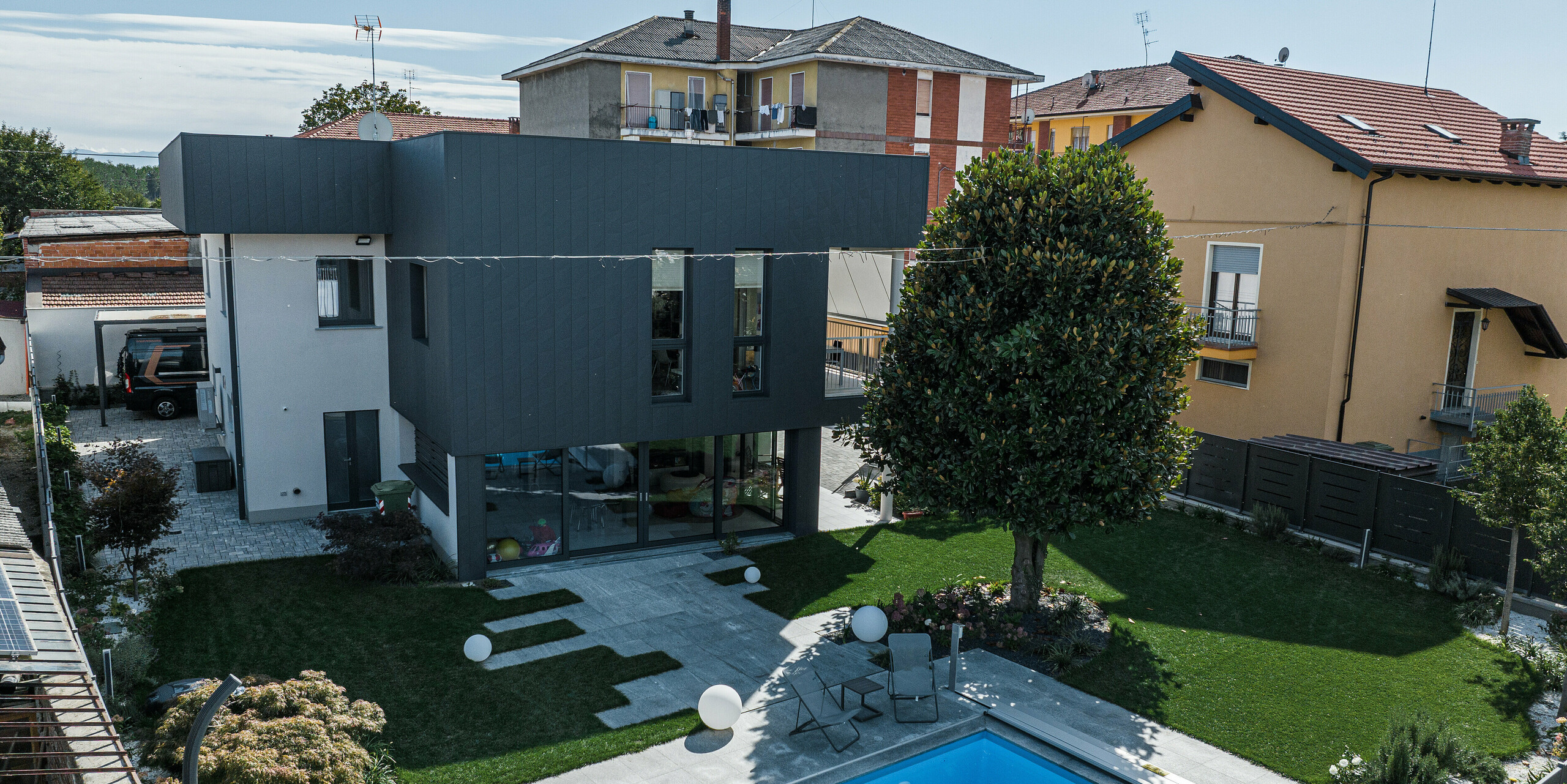 Modernes Einfamilienhaus mit einer hochwertigen PREFA Aluminiumfassade. Das Haus mit den Siding.X Fassadenpaneelen in der Farbe P.10 Dunkelgrau zeigt eine harmonische Kombination aus Funktionalität und Ästhetik, ergänzt durch einen sorgfältig gestalteten Garten mit Pool. Diese Ansicht hebt das elegante, matte Erscheinungsbild der Fassade hervor, die nicht nur optisch ansprechend, sondern auch UV-, farb- und witterungsbeständig ist. Ideal für eine nachhaltige und stilvolle Gebäudehülle.