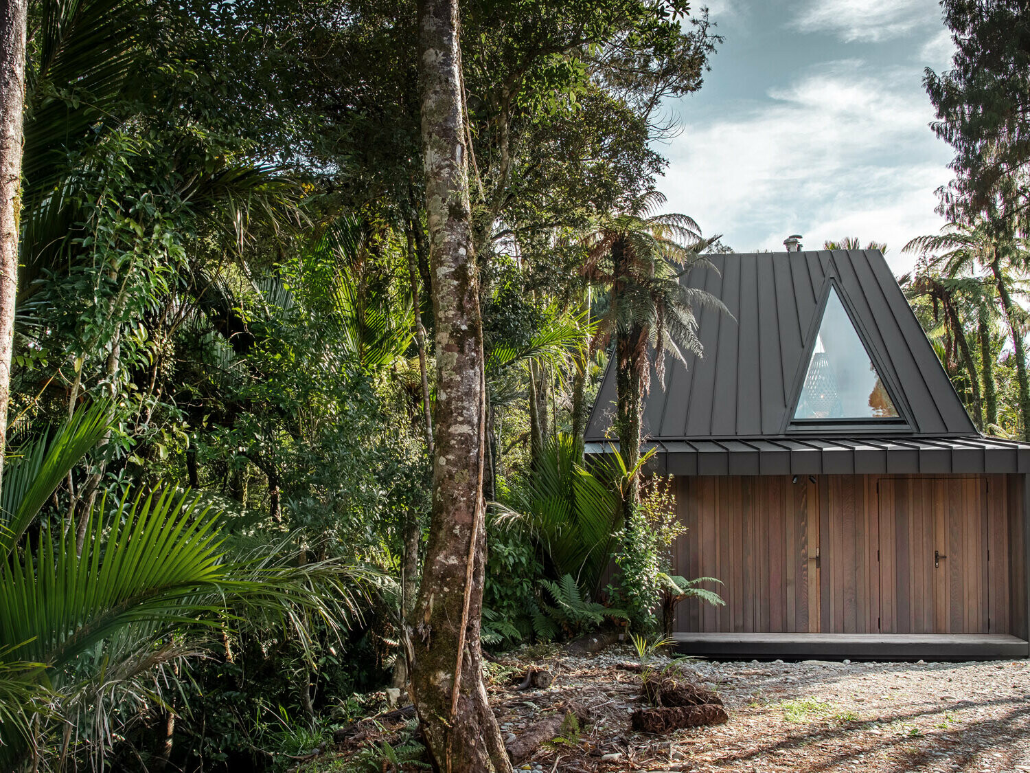Side view of the bivouac realised by Fabric with the striking, black-grey Prefalz roof hidden among nīkau palms.