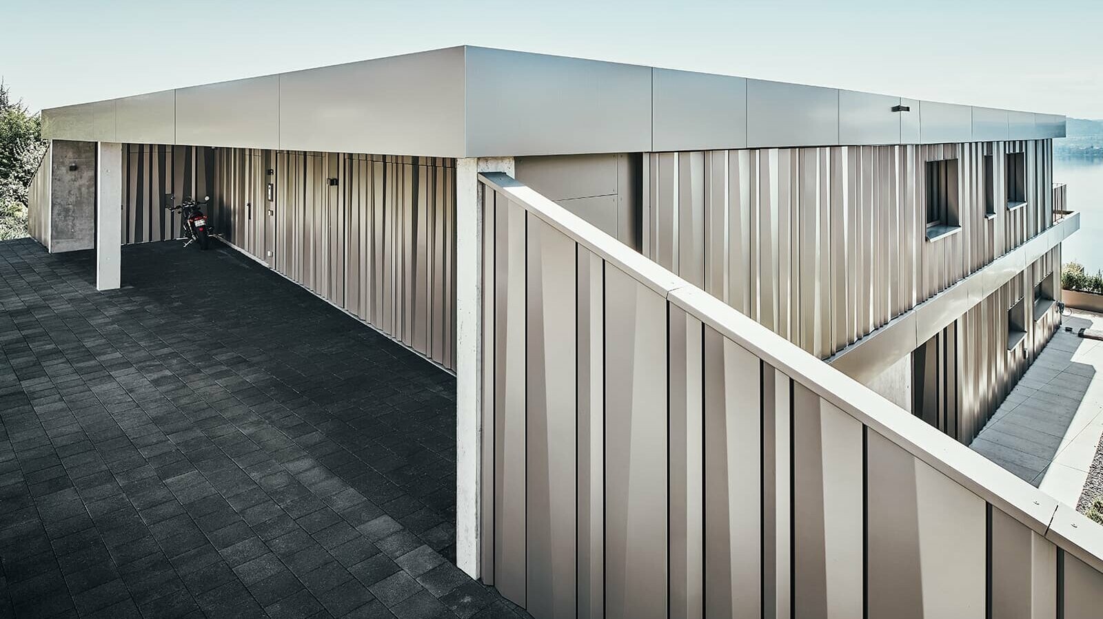 Lateral view of the detached house. The façade is covered with color aluminium coils in bronze. A motorbike is parked by the house.