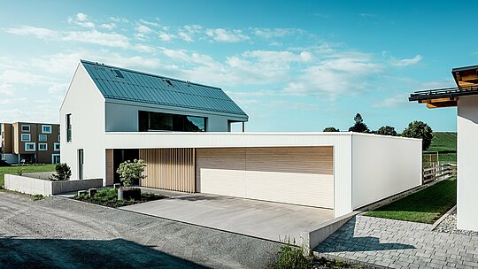 Das Bild zeigt das moderne Einfamilienhaus in Freistadt, das sich durch seine schlichte, geometrische Form und die Kombination von reinweißer PREFALZ-Aluminium-Fassade und Holzvertäfelungen auszeichnet. Die Holzverkleidung am Garagentor bietet einen warmen, natürlichen Kontrast und fügt sich perfekt in das moderne Design ein. Das Dach, ebenfalls mit PREFALZ gedeckt, sorgt für eine nahtlose, wetterfeste Oberfläche. Diese Kombination aus pflegeleichtem Aluminium und Holz schafft eine ästhetisch ansprechende und funktionale Lösung für nachhaltige Architektur. Der weitläufige, grüne Außenbereich und der klare Himmel im Hintergrund unterstreichen die moderne Lebensqualität, die dieses Haus bietet.