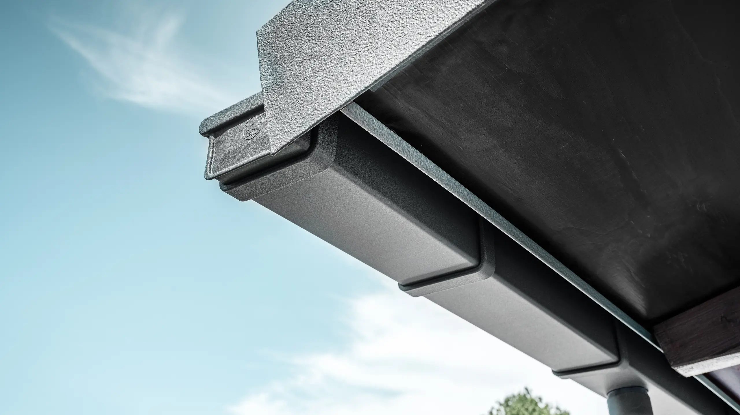 Detailaufnahme einer eckigen PREFA Kastenrinne in Anthrazit aus Aluminium mit strukturiertem Abschlussblech, montiert an der Dachkante eines Gebäudes vor blauem Himmel mit leichter Bewölkung.