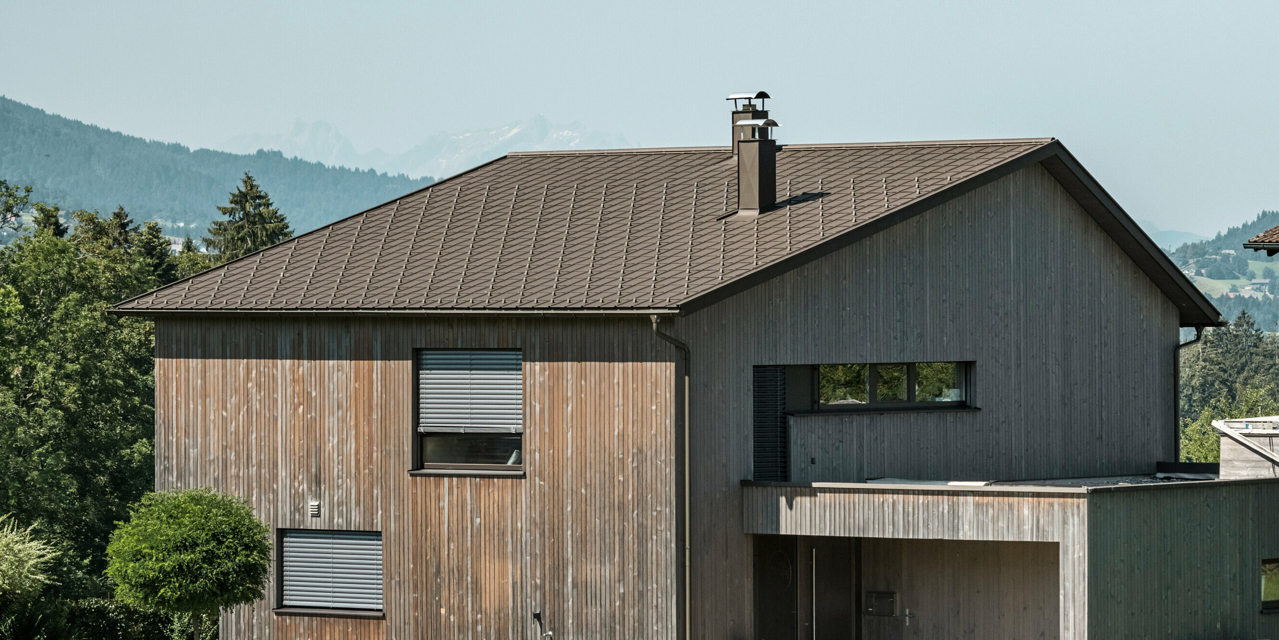 Side view of a contemporary detached house in Krumbach with a combination of a natural wooden façade and a long-lasting PREFA aluminium roof in P.10 brown. The harmonious colour scheme shows the elegant integration of modern design and traditional materials, with the PREFA roof tiles providing an aesthetic and functional roof design.