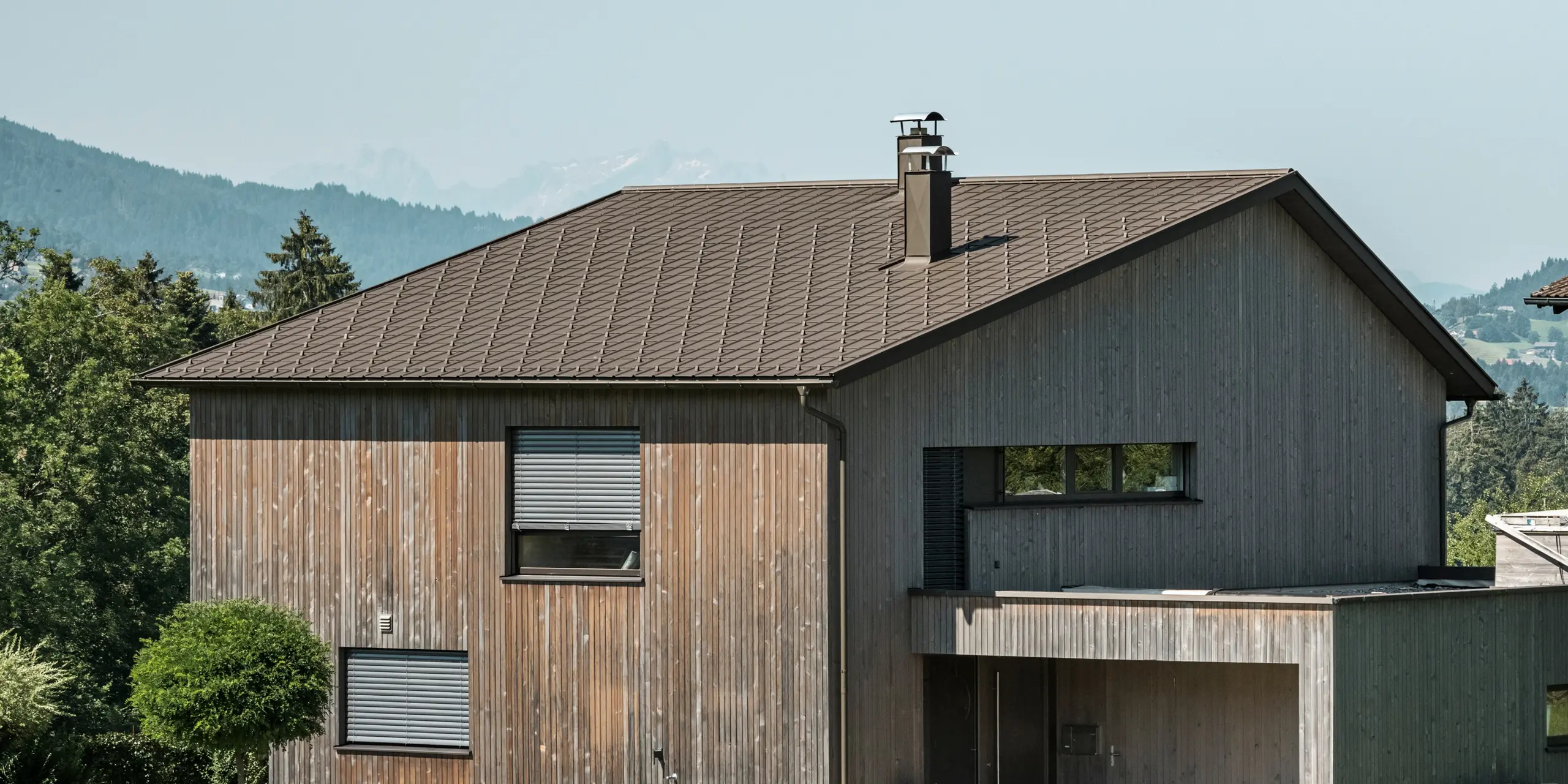 Side view of a contemporary detached house in Krumbach with a combination of a natural wooden façade and a long-lasting PREFA aluminium roof in P.10 brown. The harmonious colour scheme shows the elegant integration of modern design and traditional materials, with the PREFA roof tiles providing an aesthetic and functional roof design.