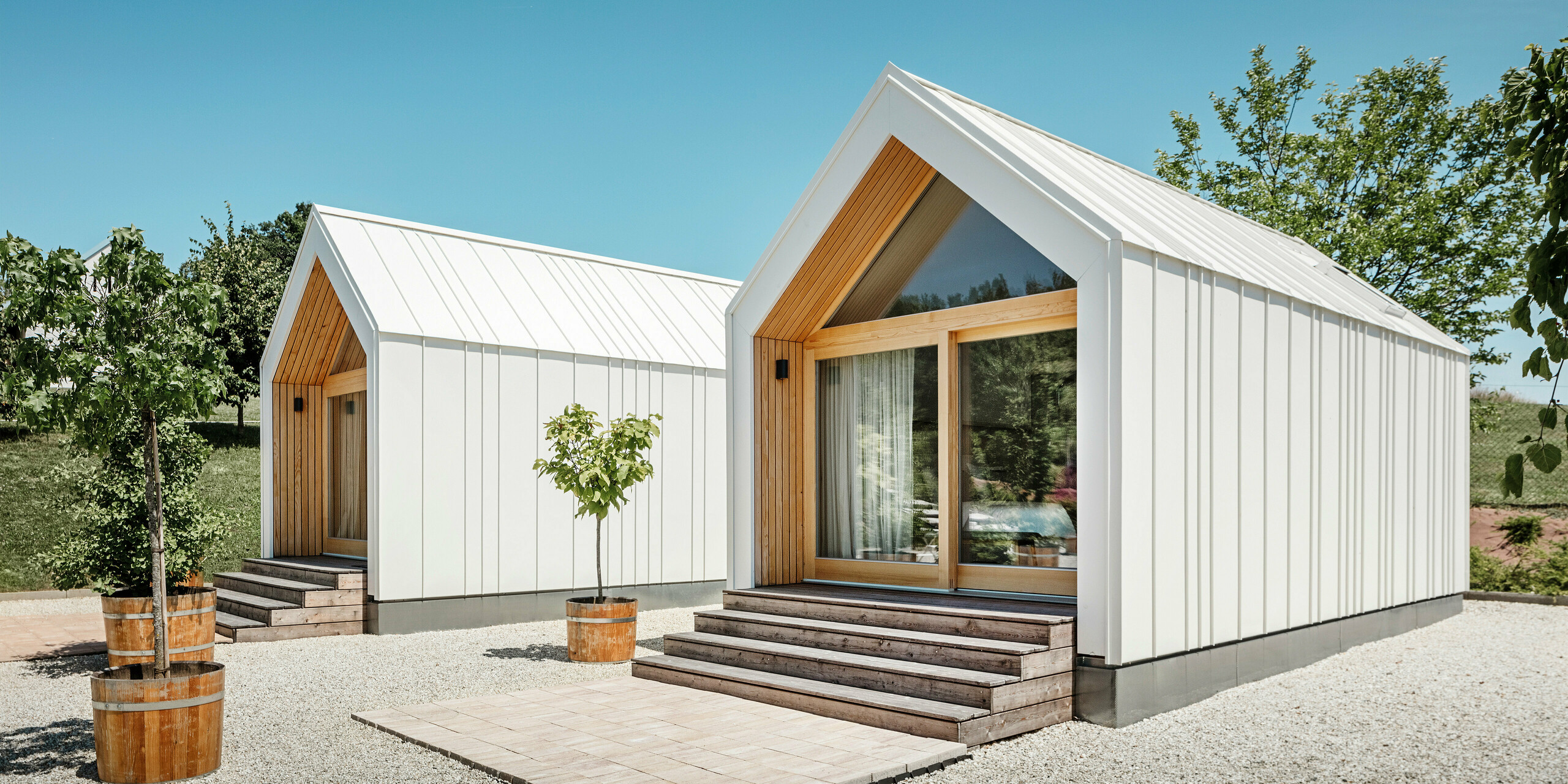 Two white tiny houses in the holiday idyll 'Pri Momi' in Kančevci, Slovenia, designed with PREFALZ roof and façade cladding in P.10 pure white and natural wood. The minimalist architecture of the buildings is emphasised by the elegant material mix of aluminium and wood. The large glass fronts at the entrances as well as the wooden steps and surrounding trees create an inviting and harmonious atmosphere. The exclusive accommodation is located in the middle of a picturesque landscape and is used especially for celebrations.