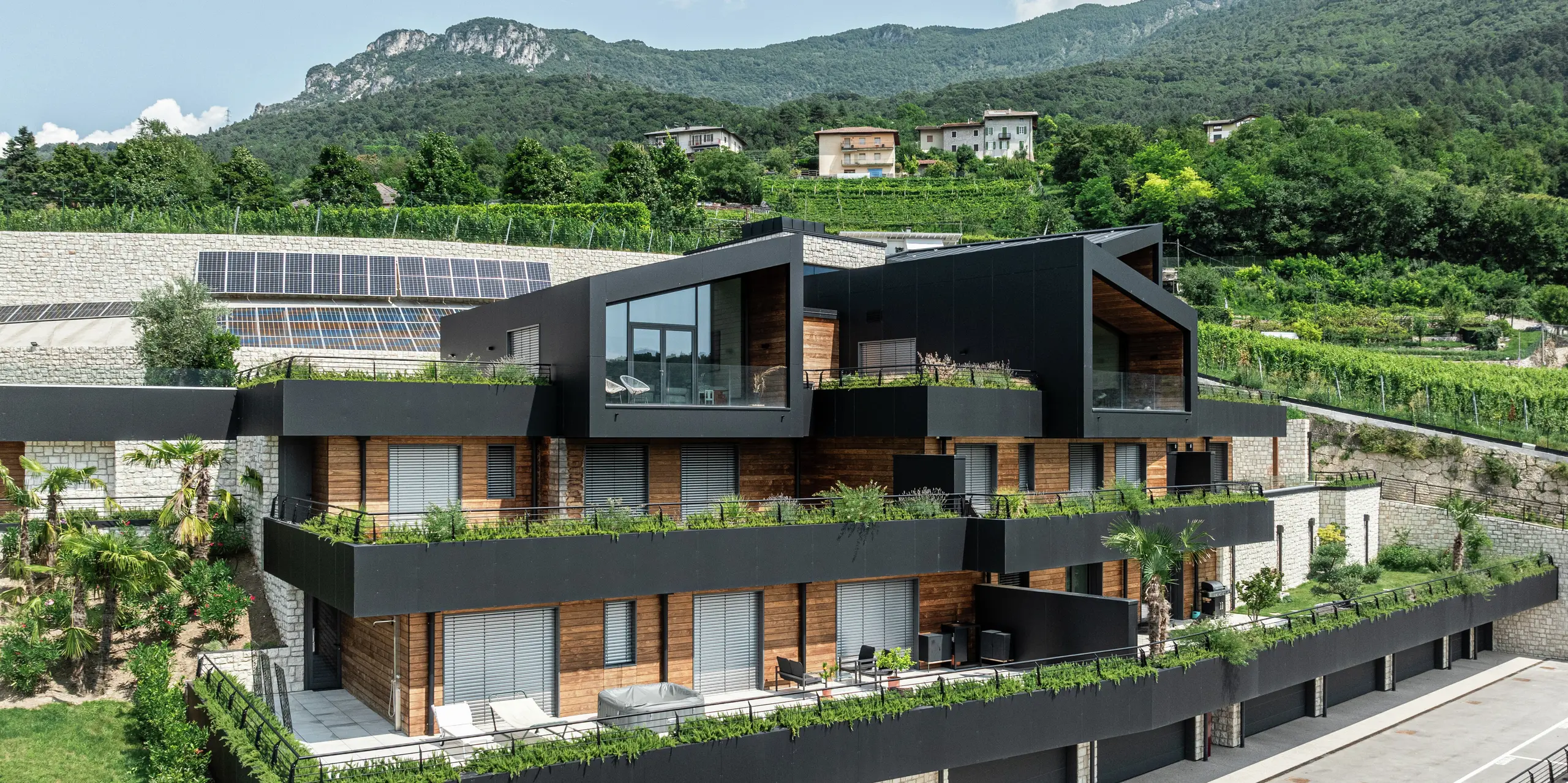 Moderne Wohnanlage in Hanglage mit nachhaltigem Design: Die Kombination aus Naturstein, Holz und der hochwertigen PREFA Aluminiumfassade in P.10 Schwarz verleiht dem Gebäude eine elegante und zeitlose Optik. Das PREFALZ Dach in P.10 Schwarz bietet optimalen Schutz und Langlebigkeit. Großzügige Terrassen, begrünte Dächer und Photovoltaikanlagen unterstreichen das nachhaltige Wohnkonzept. Die harmonische Einbettung in die Landschaft sowie die hochwertigen und pflegeleichten PREFA Materialien garantieren eine stilvolle und witterungsbeständige Architektur mit höchstem Wohnkomfort.