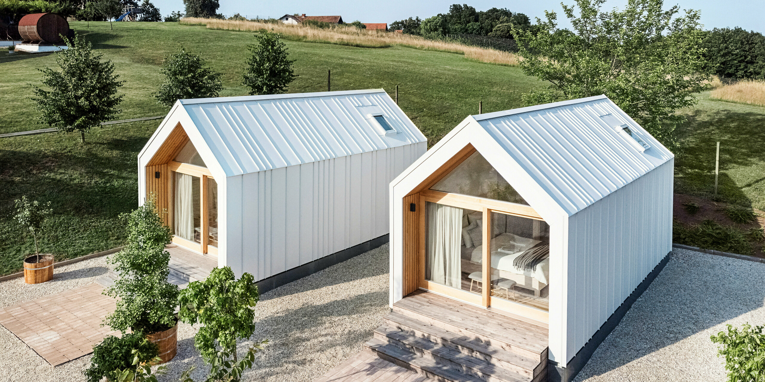 View of the front of the two tiny houses in the 'Pri Momi' holiday complex in Kančevci, Slovenia. The micro houses were built using a timber frame construction and finished with a robust PREFALZ roof and façade cladding in P.10 Pure White. The modern, minimalist buildings combine a durable yet aesthetic aluminium shell with large glass fronts and natural wood, embedded in a green landscape. The generous wooden steps lead up to the entrances, creating a cosy and inviting atmosphere.