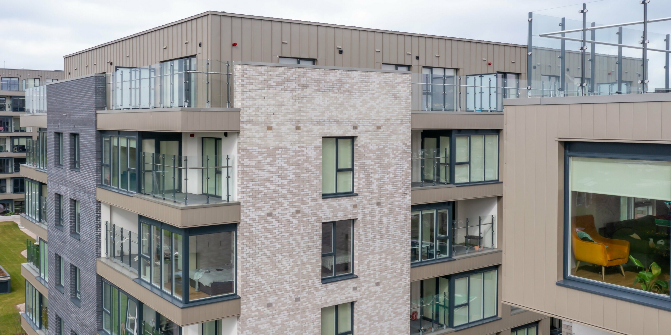 Drone footage of the Palmers Gate residential complex in Palmerstown, Dublin, Ireland - detailed view of a residential building with bronze-coloured PREFA cladding and PREFALZ façade system. The façade combines aluminium cladding with brick elements and large glass windows. The balconies are equipped with glass balustrades and offer modern living quality. The building complex consists of several blocks of flats with approx. 1,700 m² of PREFA sidings and approx. 1,200 m² of PREFALZ façade system, which are known for their durability and aesthetic design.