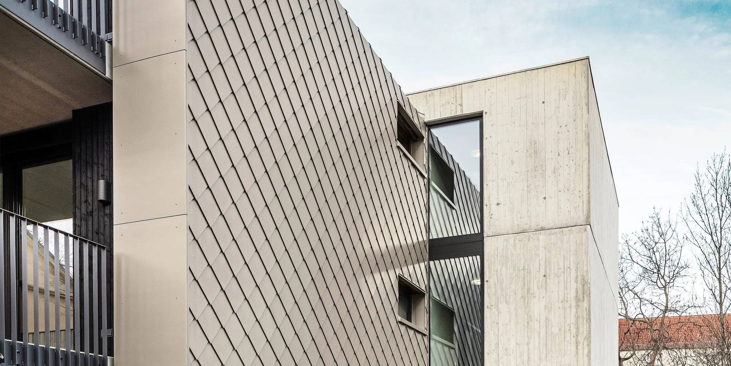 Zugangsbereich zu einem Mehrfamilienhaus in der Chemnitzer Straße im Bezirk Plauen, Dresden. Das Gebäude wird geschützt von einem robusten PREFA Dach- und Fassadensystem aus Aluminium. Die hochwertigen PREFA Dach- und Wandrauten 29 × 29 in Bronze sorgen für eine elegante Optik und langlebigen Schutz. Das Bild zeigt den Eingangsbereich, der von der eleganten Rautenstruktur und einer Betonwand flankiert wird. Die architektonische Gestaltung kombiniert robuste Materialien mit klaren Linien und modernen Elementen, was dem Gebäude eine zeitgemäße und stilvolle Optik verleiht.
