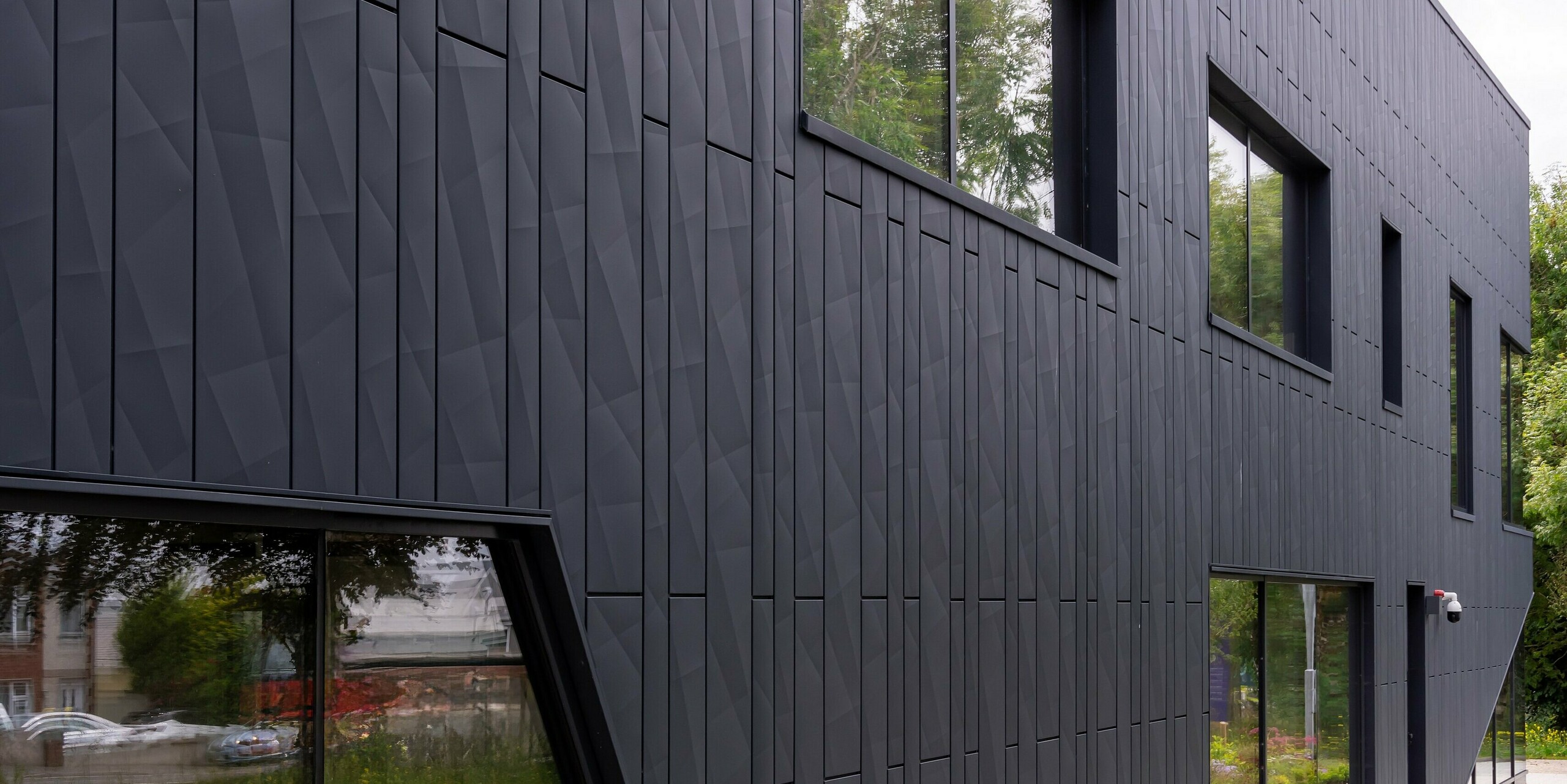 Rear view of the CREW building in Galway, Ireland, with a detailed view of the PREFA Siding.X façade panels in P.10 anthracite. The geometrically structured aluminium cladding gives the façade a modern and dynamic look. The building's individual window areas and clean lines complement the minimalist design, while the surrounding greenery completes the overall picture.