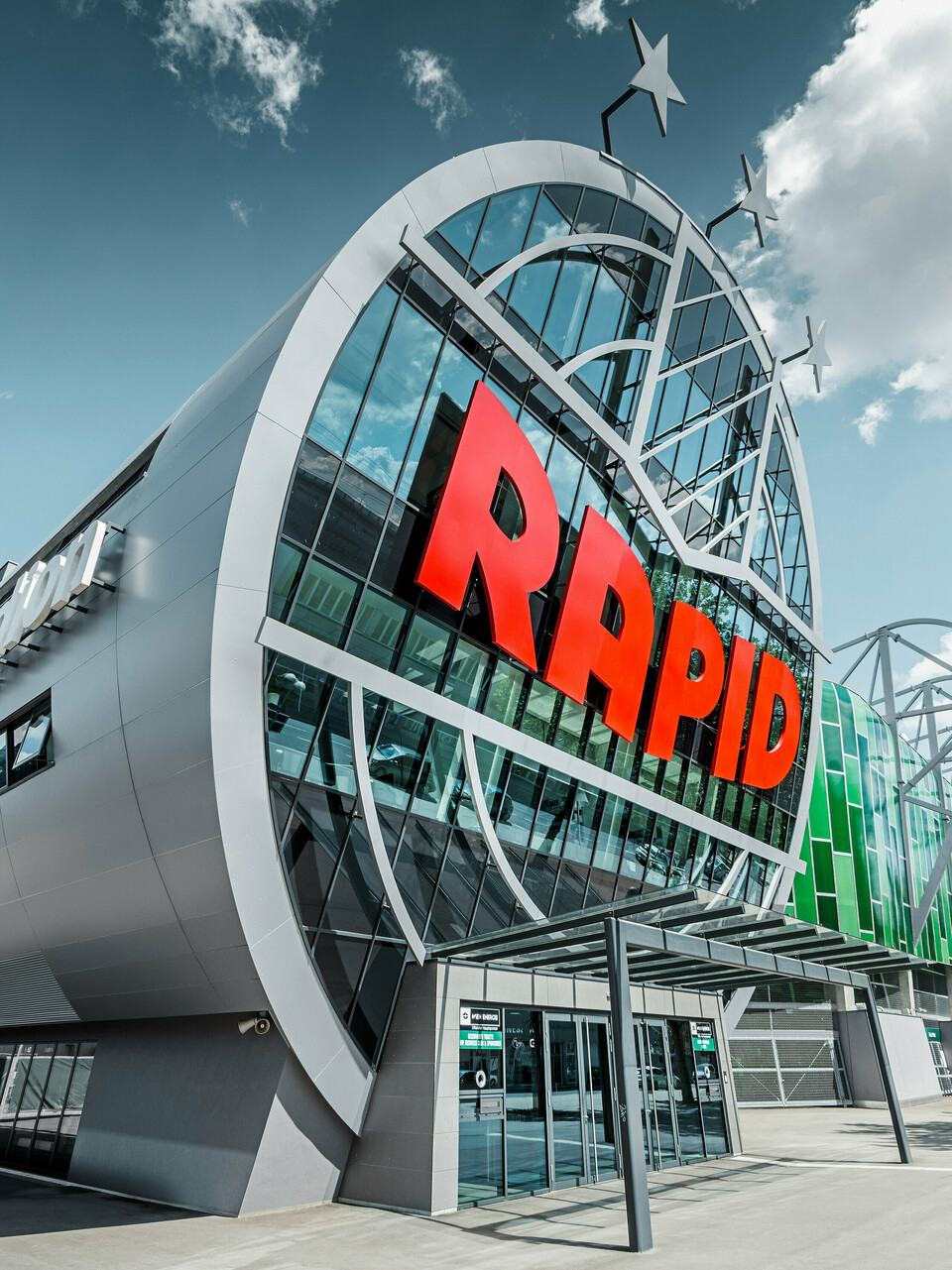 Ansicht des Allianz Stadions von SK Rapid Wien mit auffälliger PREFA Fassade in Form der Aluminium Verbundplatte in Silbermetallic. Die Gebäudehülle strahlt Dynamik und Moderne aus. Der Eingangsbereich, gekrönt mit dem leuchtend roten RAPID-Schriftzug, lädt ein in das Herz des Fußballs, eingefasst von einer futuristischen und zugleich eleganten Fassadenstruktur, die Sportarchitektur auf ein neues Niveau hebt.