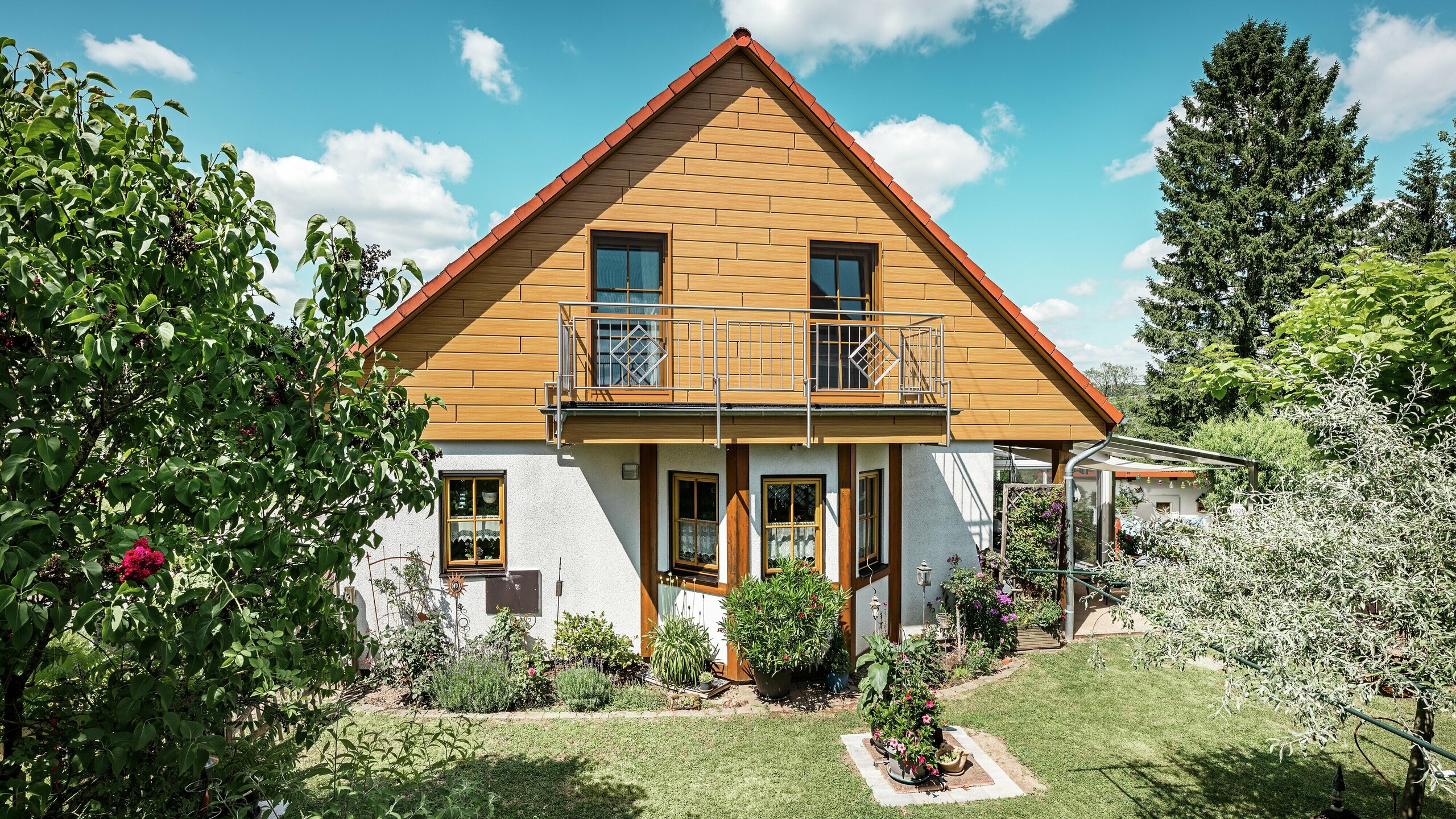 In this photo, the renovated gable end of a detached house in Blieskastel shines in a warm natural oak tone thanks to PREFA Sidings. The high-quality aluminium façade, with its realistic wood-effect finish, creates a striking visual feature and blends impressively with the red tiled roof and the lush garden. Surrounded by flowers, shrubs and lush greenery, the house looks inviting and well-maintained. The PREFA surface combines a natural look with functional benefits such as high weather resistance, stability and virtually maintenance-free sustainability – an aesthetic highlight with sustainable added value.