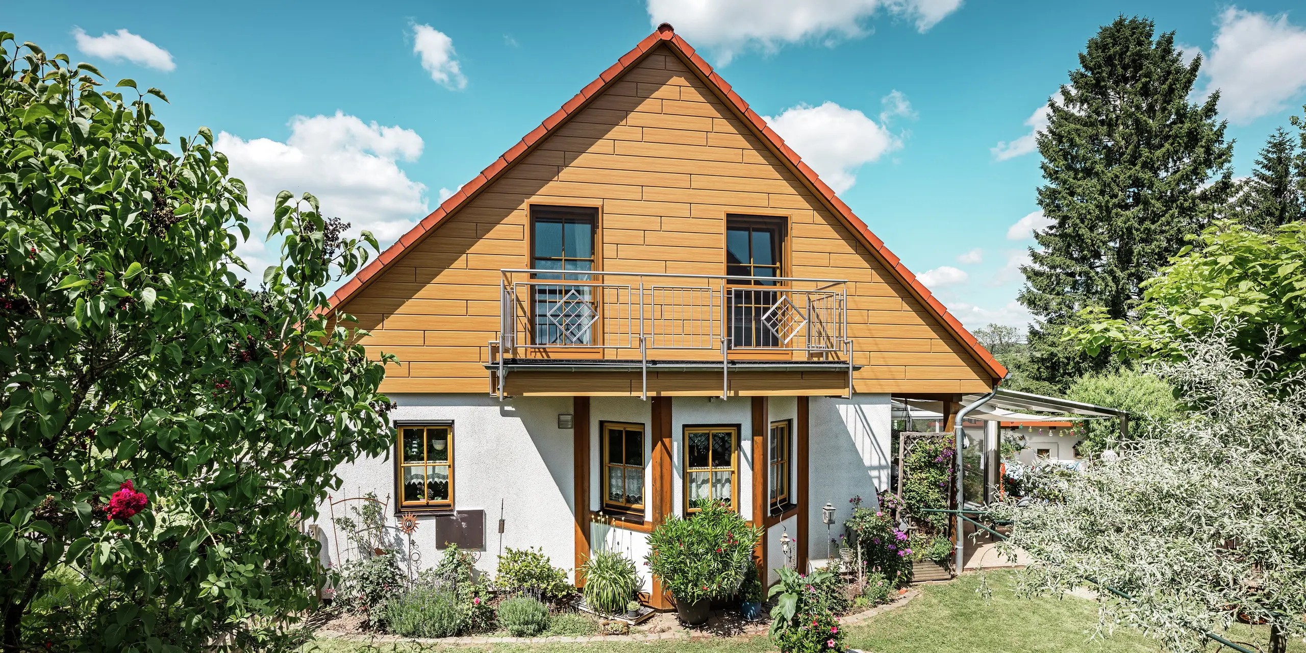 In this photo, the renovated gable end of a detached house in Blieskastel shines in a warm natural oak tone thanks to PREFA Sidings. The high-quality aluminium façade, with its realistic wood-effect finish, creates a striking visual feature and blends impressively with the red tiled roof and the lush garden. Surrounded by flowers, shrubs and lush greenery, the house looks inviting and well-maintained. The PREFA surface combines a natural look with functional benefits such as high weather resistance, stability and virtually maintenance-free sustainability – an aesthetic highlight with sustainable added value.