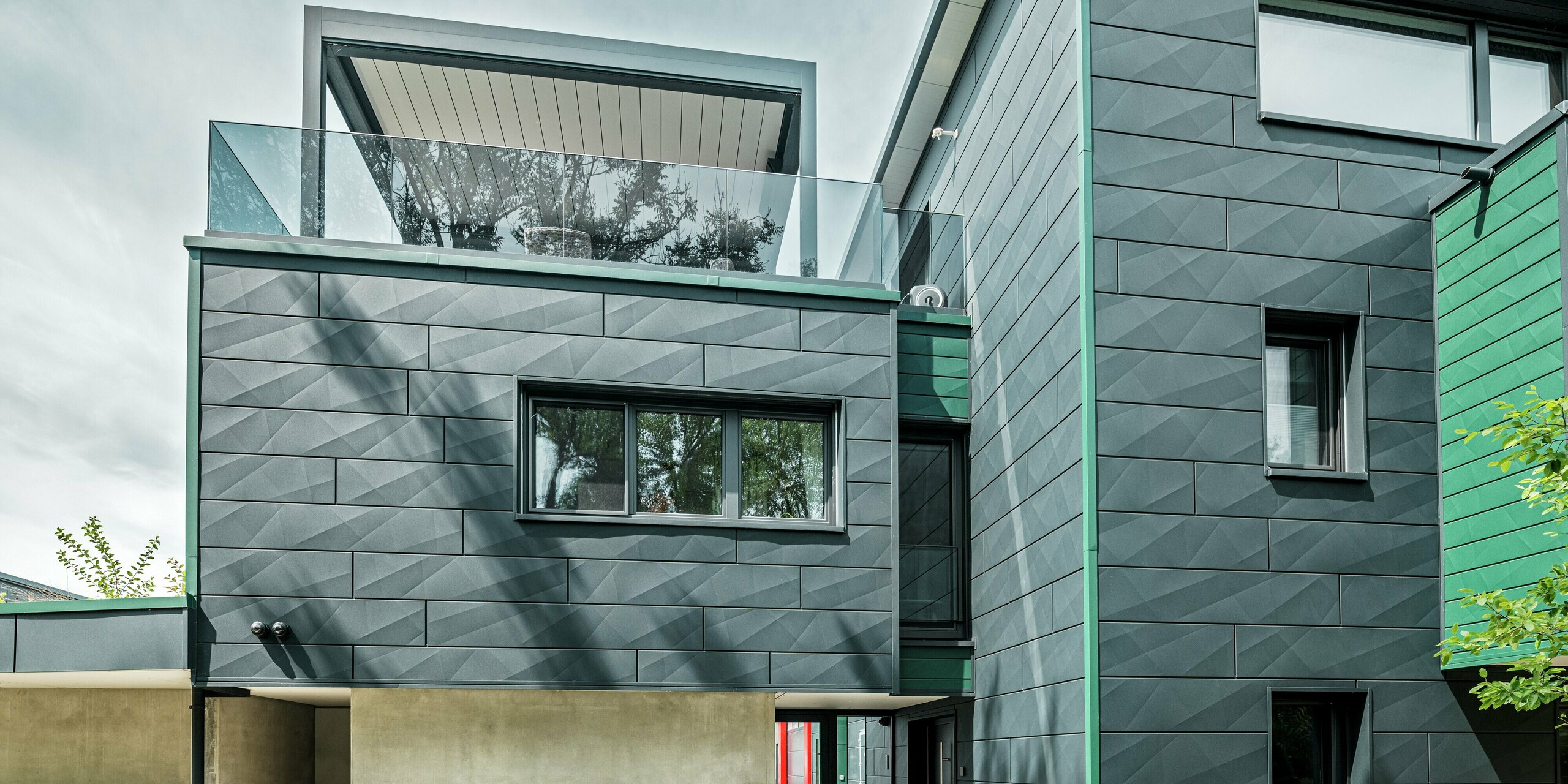 Das moderne Einfamilienhaus in Türkheim beeindruckt mit seiner einzigartigen Fassade aus PREFA Siding X-Paneelen in den Farben P.10 Anthrazit und P.10 Moosgrün. Die Längs- und Querkantungen der Aluminiumpaneele verleihen dem Gebäude eine dynamische Optik. Die Perspektive zeigt das Haus von der Gartenseite aus mit Blick auf den Eingangsbereich. Über dem Eingangsbereich ist eine Dachterrasse mit einer modernen Pergola zu erkennen.