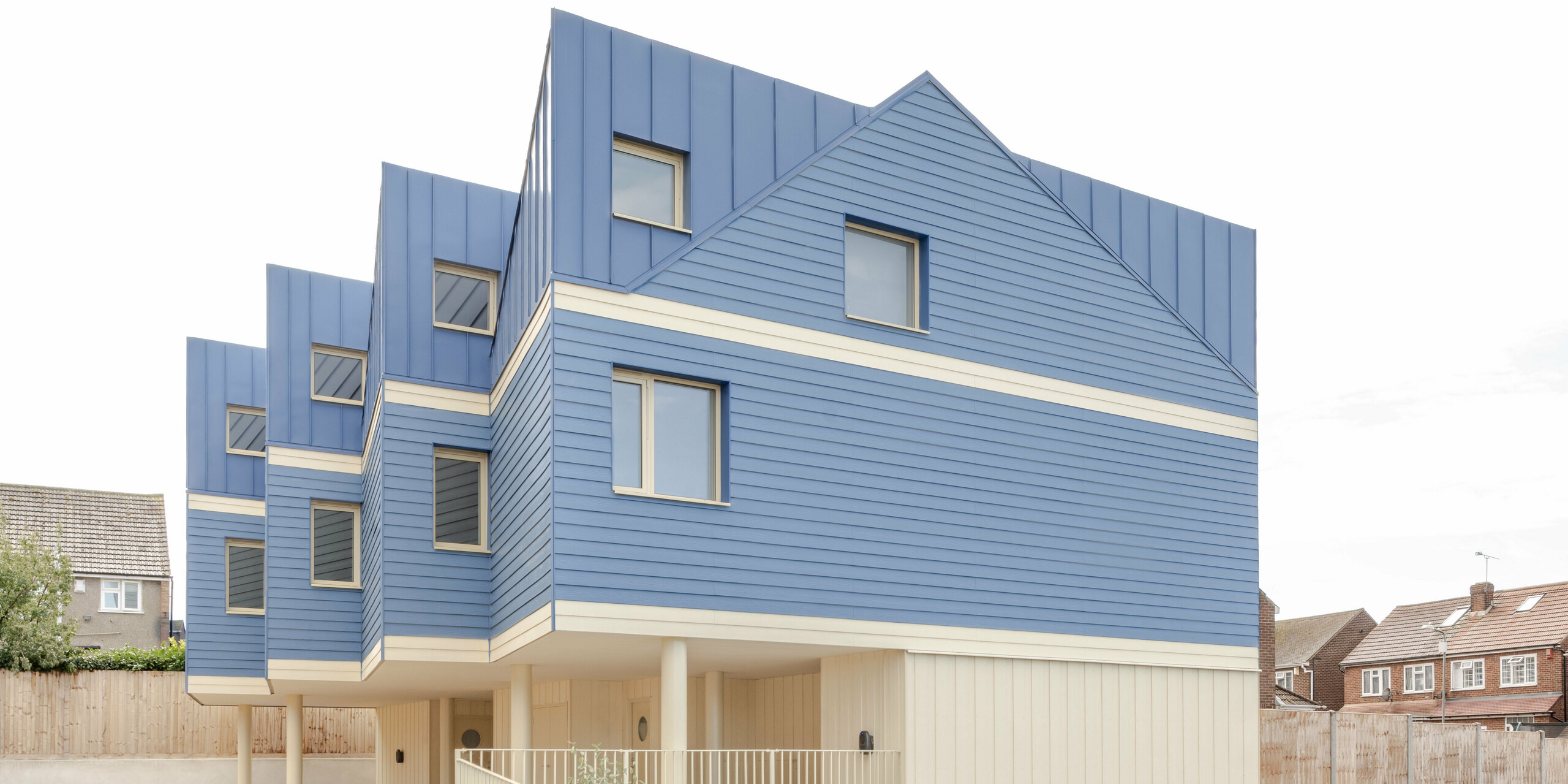 The image shows the striking front of the Goldsmith Mews terraced housing complex with its eye-catching FALZONAL standing seam cladding in dusty blue. The precise, vertical folded panels on the roof and upper floor blend harmoniously with the horizontal aluminium profiles, creating a lively, rhythmic façade. The light-coloured lower areas and the elevated construction lend the building a sense of lightness. The durable, colour-stable PREFA aluminium offers reliable weather protection, high dimensional stability and enables a modern, clear architecture with strong recognition value.