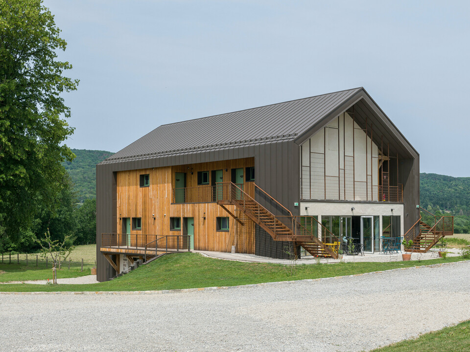 Eckansicht eines zeitgenössischen zweigeschossigen Hotels mit einer Hülle aus PREFA Aluminium und naturbelassener Holzfassade. Das Dach sowie Teile der Fassade wurden mit PREFALZ in P.10 Braun verkleidet. Hervorstechende Merkmale des Gebäudes sind große Fensterflächen sowie eine außenliegende Treppe, die zum oberen Stockwerk führt. Zudem verfügt das Hôtel en Tilleul über eine Terrasse im Erdgeschoss. Das Gebäude steht in einer idyllischen ländlichen Umgebung mit Blick auf sanfte Hügel. Am unteren Bildrand ist die Zufahrtsstraße aus Kies zu sehen. 