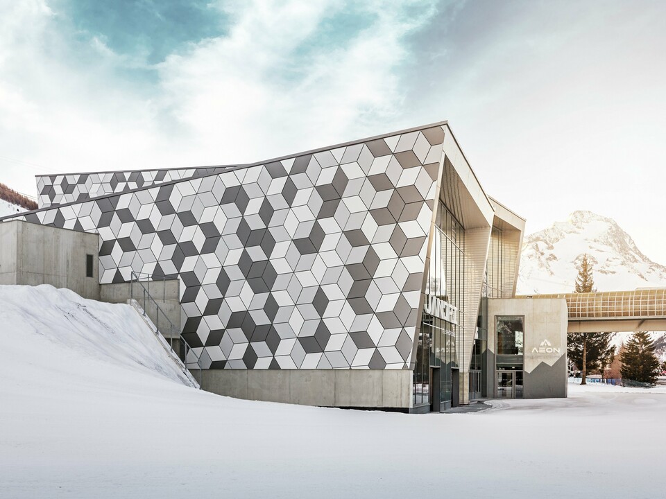 Die Talstation in Les Deux Alpes beeindruckt mit einer außergewöhnlichen Fassade aus PREFABOND Aluminium-Verbundplatten in einem modernen Rautenmuster. Die Kombination aus Weiß- und Grautönen bildet ein markantes Design, das perfekt mit der verschneiten Berglandschaft harmoniert. Die langlebige, wetterbeständige Fassade garantiert auch unter extremen Bergbedingungen eine ästhetische und funktionale Lösung. Die strahlende Sonne im Hintergrund schafft einen majestätischen Rahmen für diese Szenerie.