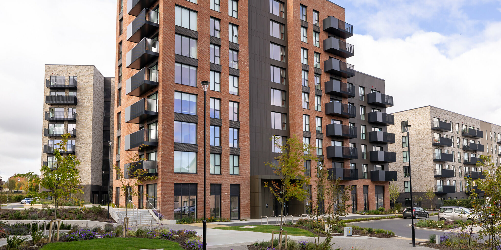 Front view of the Citywest Apartments in Dublin. The residential building combines a traditional brick façade with a modern design of vertically laid PREFA sidings in P.10 brown. Spacious balconies and large windows emphasise the modern architecture. The weather-resistant aluminium façade offers a stylish and durable solution that blends harmoniously into the urban environment with its well-kept green areas.  Translated with DeepL.com (free version)