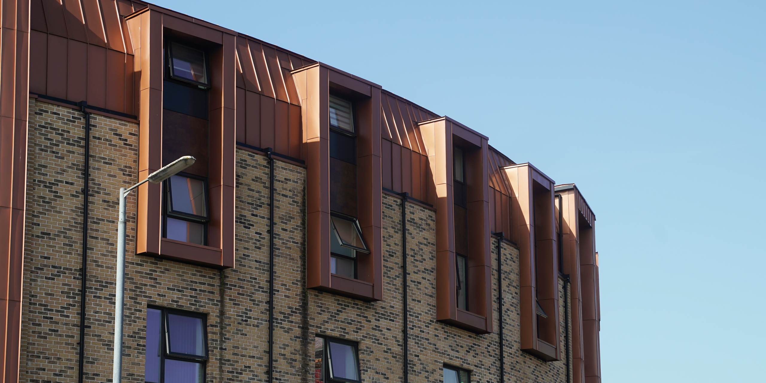 The façade of a residential development on Nethan Street in Glasgow, accentuated by the tasteful use of FALZONAL in New Copper. The PREFA aluminium product gives the building envelope a vibrant and inviting appearance through its reflection of sunlight. The combination of the traditional brick structure with the modern, copper-coloured accents of the FALZONAL conveys an elegant symbiosis of old and new and highlights the architecture in this urban context.