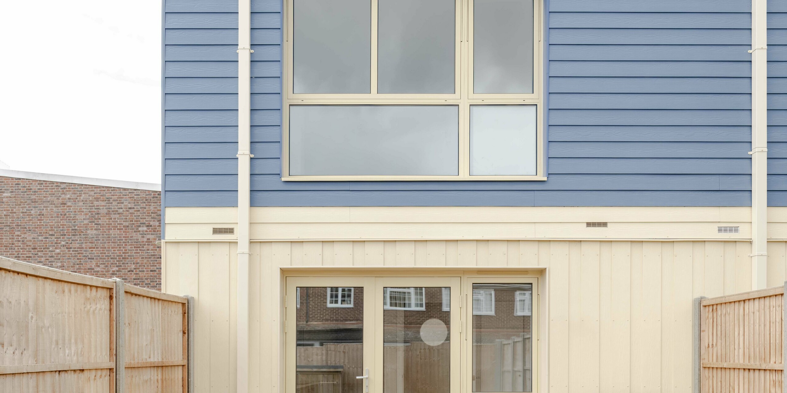 The photo shows the reverse of a Goldsmith Mews terraced house with striking FALZONAL standing seam cladding in dusty blue. The clearly structured vertical aluminium folds in the upper section meet the horizontal panels of the middle storey, creating a strongly structured façade. The light-coloured lower sections with large patio doors open up the architecture to the garden. The high-quality PREFA aluminium offers high colour and shape stability, lasting weather protection and enables a modern, precisely designed building envelope with clear recognition.