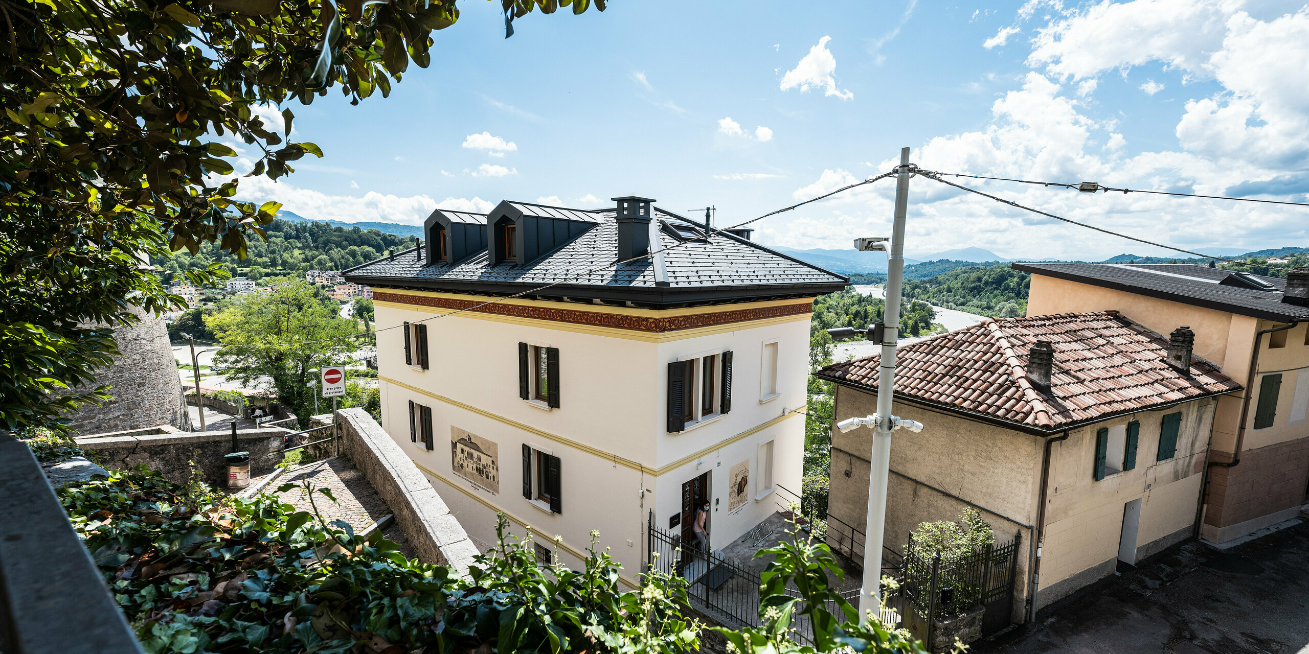 Panoramaansicht eines sanierten, historischen Wohngebäudes in Belluno, Italien, eingebettet in ein städtisches Umfeld mit Blick auf grüne Hügel. Die elegante Fassade im klassischen Stil des frühen 20. Jahrhunderts wird durch ein PREFA Aluminiumdach mit anthrazitfarbenen Dachschindeln ergänzt, die für eine zeitgemäße, nachhaltige Lösung sorgen. Die sanfte Integration der PREFALZ Dachgauben und Kamine in P.10 Anthrazit bietet eine ansprechende Optik und spiegelt das Engagement für den Erhalt historischer Architektur wider.