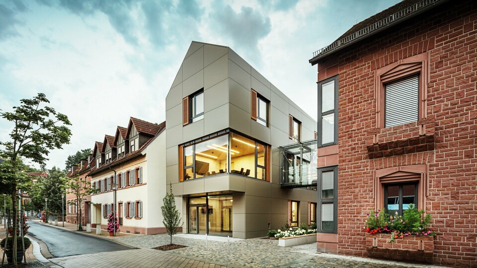 The image shows the modern town hall in Alzenau, which is clad in aluminium composite panels from PREFA in a warm bronze tone. The façade is interrupted by large windows that offer a view of the interior, which is illuminated by warm light. The glass walkway on the right-hand side of the picture connects the building to a neighbouring traditional brick building, creating an exciting contrast between modern and historic architecture.