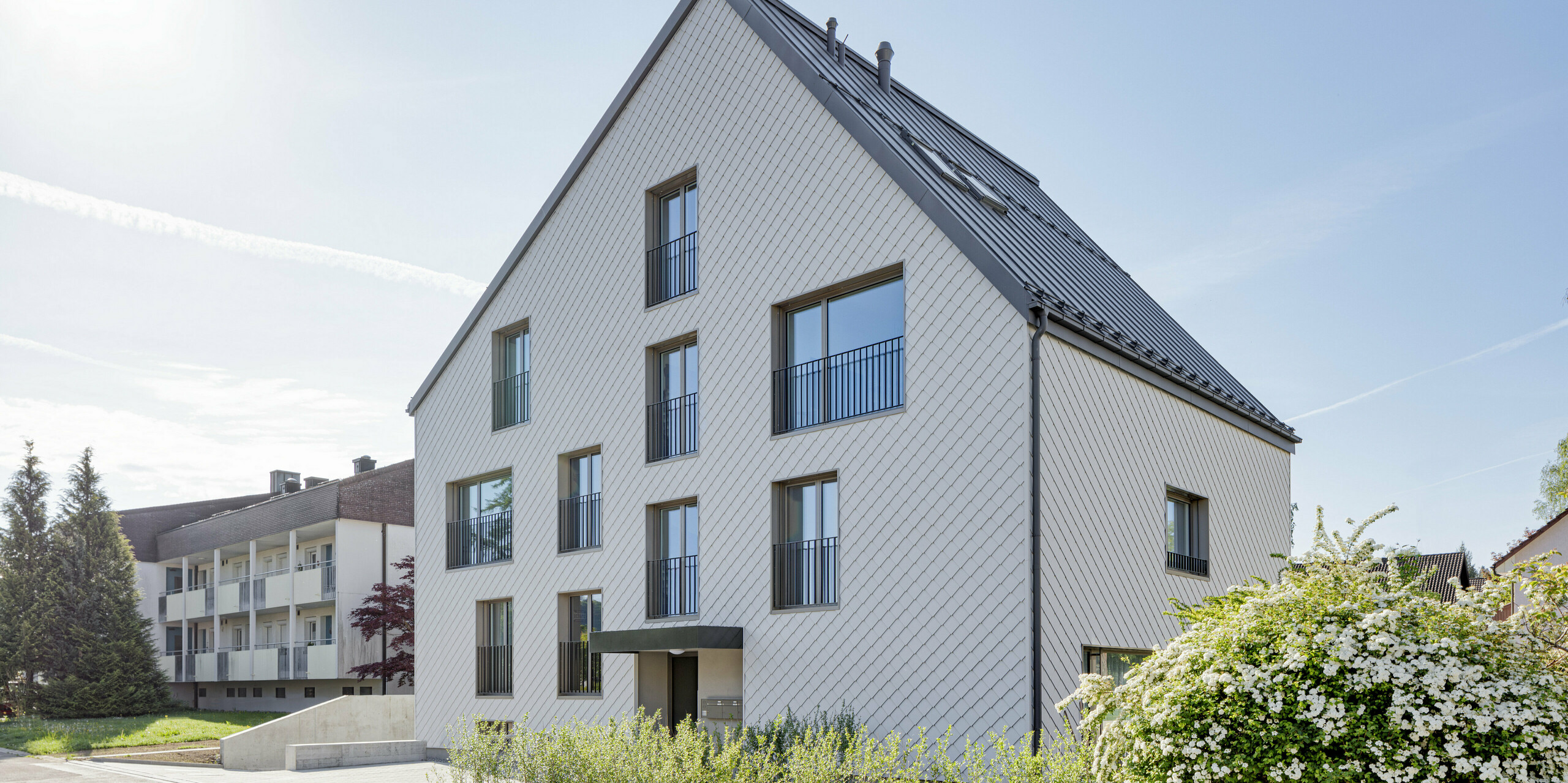 Modernes Mehrfamilienhaus in der Schweiz mit einem langlebigen PREFA Dach aus PREFALZ in P.10 Dunkelgrau und einer Fassade, verkleidet mit PREFA Wandrauten 20x20 in P.10 Prefaweiß. Das Objekt bietet eine zeitgemäße Architektur, die durch die Kombination von traditionellem Satteldach und minimalistischen Fassadenelementen besticht. Die klare Linienführung und das strukturierte Erscheinungsbild der Wandrauten sind harmonisch in eine gepflegte Umgebung mit üppigem Blütenstrauch integriert und zeigen die Vielseitigkeit von Aluminium als Baumaterial.