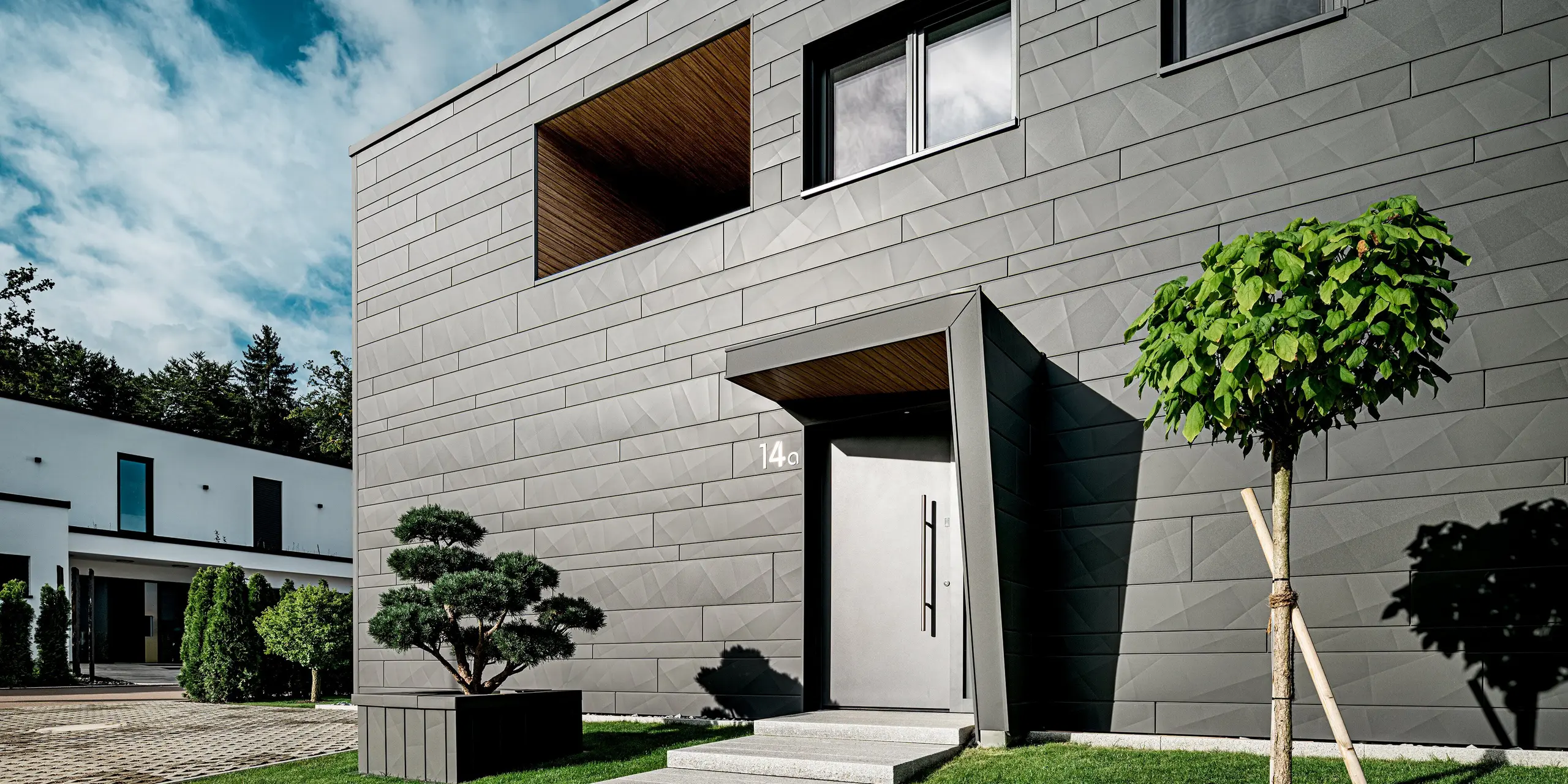 Das moderne Einfamilienhaus in Heidenheim präsentiert sich mit einer markanten PREFA Siding.X Fassade in P.10 Dunkelgrau, deren kantige Struktur das klare Design betont. Der Eingang mit tiefem, holzverkleidetem Rücksprung erzeugt einen spannenden architektonischen Fokuspunkt. Im Vordergrund setzen ein geschnittener Formbaum und ein junger Kugelbaum lebendige Akzente. Die widerstandsfähige Aluminiumfassade sowie das integrierte PREFA Quadratrohr sorgen für dauerhaften Schutz, moderne Optik und ein hochwertiges Gesamtbild.