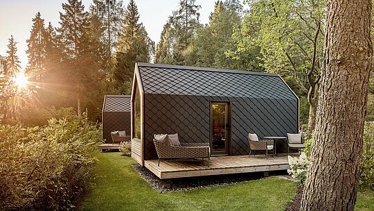 Ein Tiny House des Secret Koi Garden liegt eingebettet in eine ruhige Waldlandschaft am Natterer See, warm beleuchtet von tiefstehender Abendsonne. Dach und Fassade sind mit kleinteiligen PREFA Rauten aus Aluminium in P.10 Anthrazit verkleidet, deren strukturierte Oberfläche an Fischhaut erinnert. Die robuste, langlebige Aluminiumhülle schützt zuverlässig vor Witterung und unterstreicht die klare, zeitlose Glamping‑Architektur inmitten der Natur.